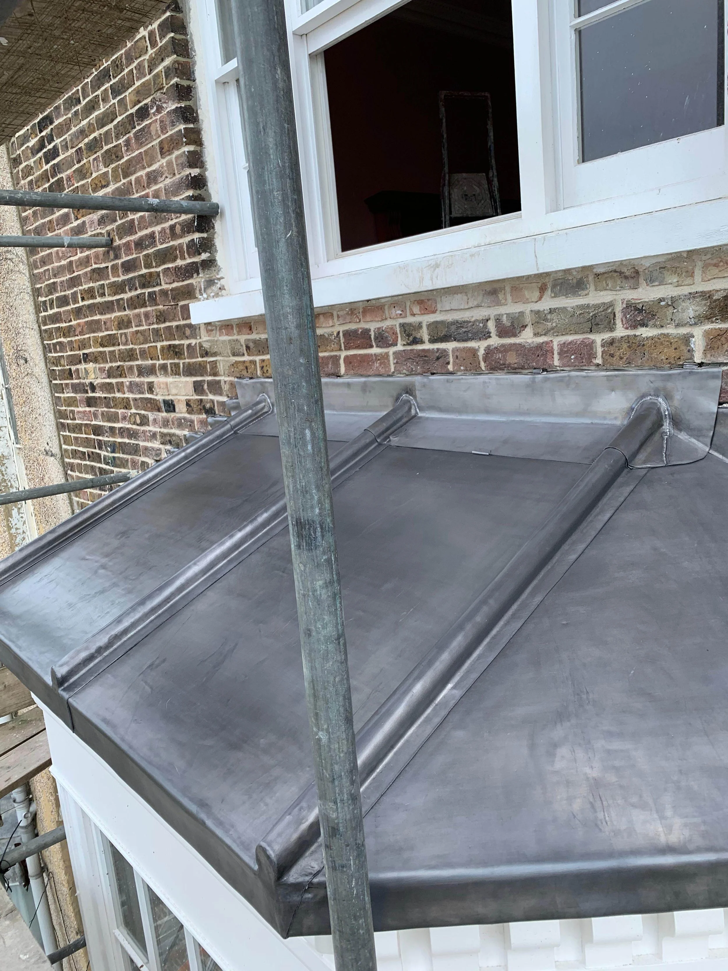 Metal flashing installed above a window on a brick building, with scaffold poles nearby.