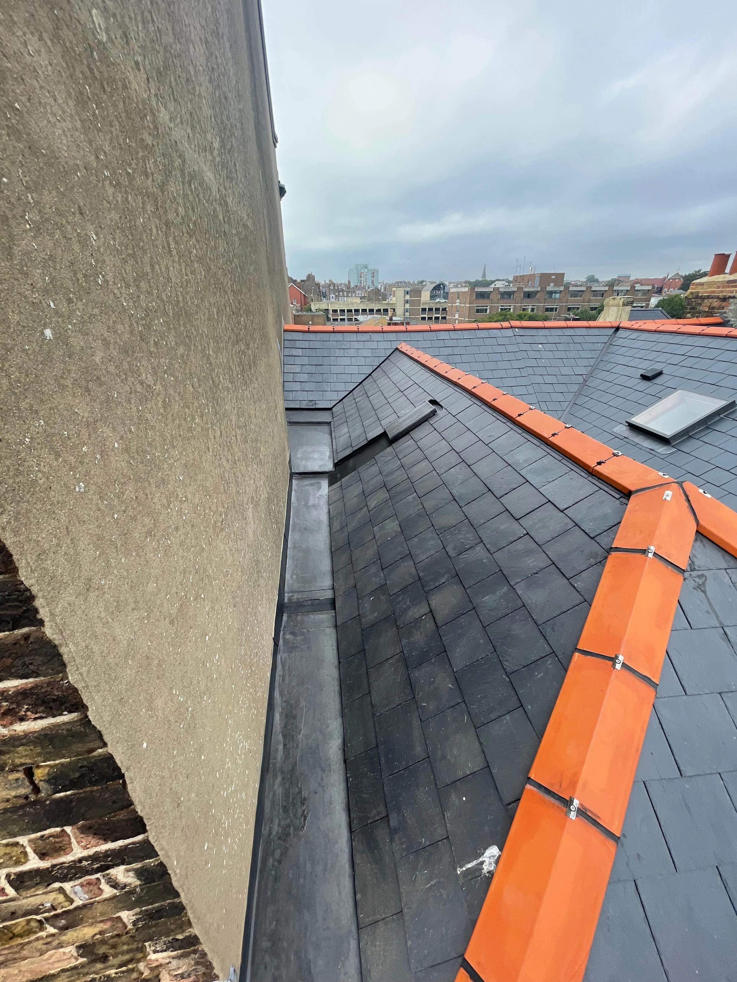 View of a rooftop with black slate tiles, orange ridge tiles, skylights, and a chimney, with an urban cityscape in the background under a cloudy sky.