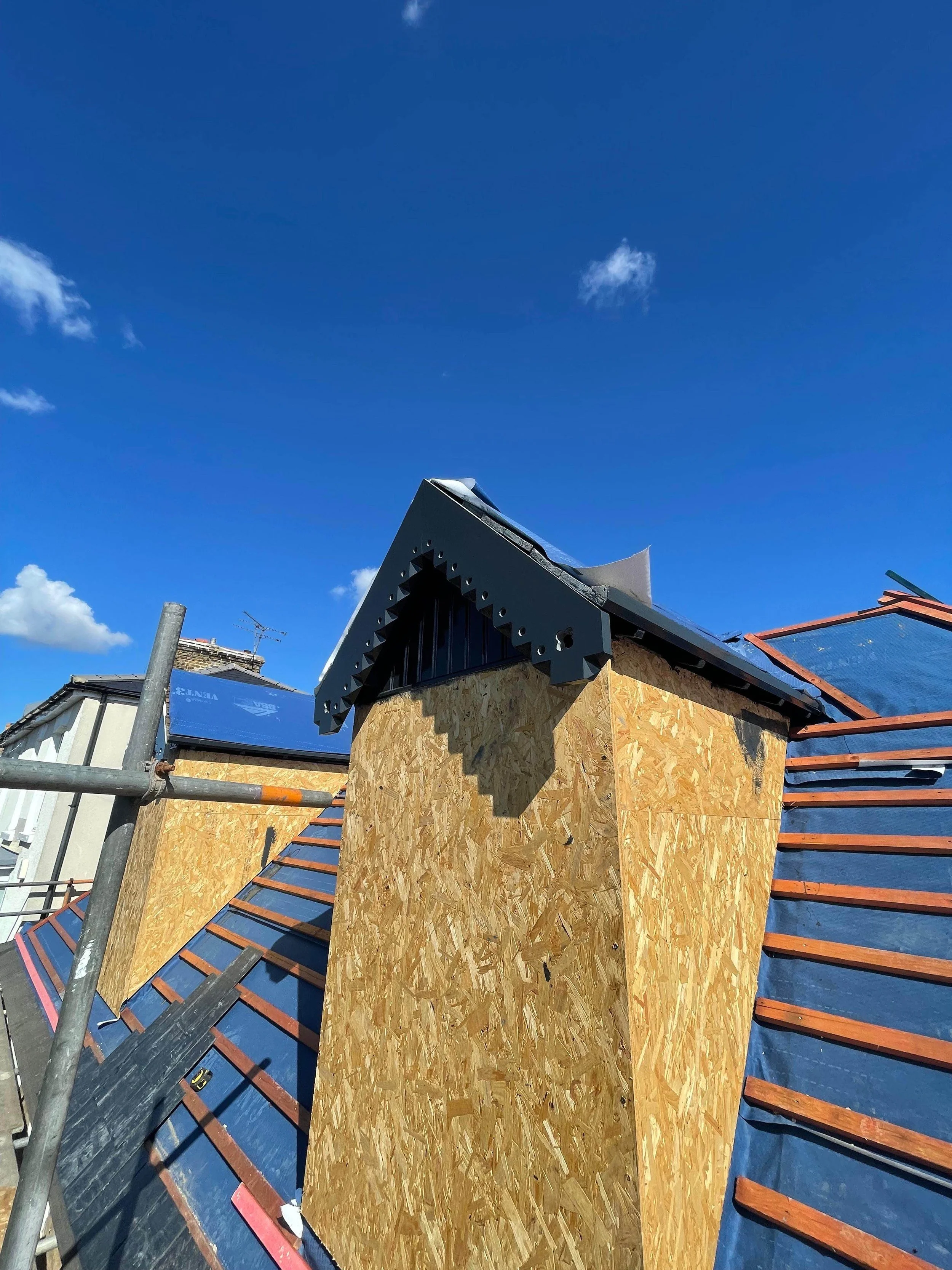 Photograph of a construction site showing a roof with blue panels and a chimney covered with plywood, under a clear blue sky with a few clouds.