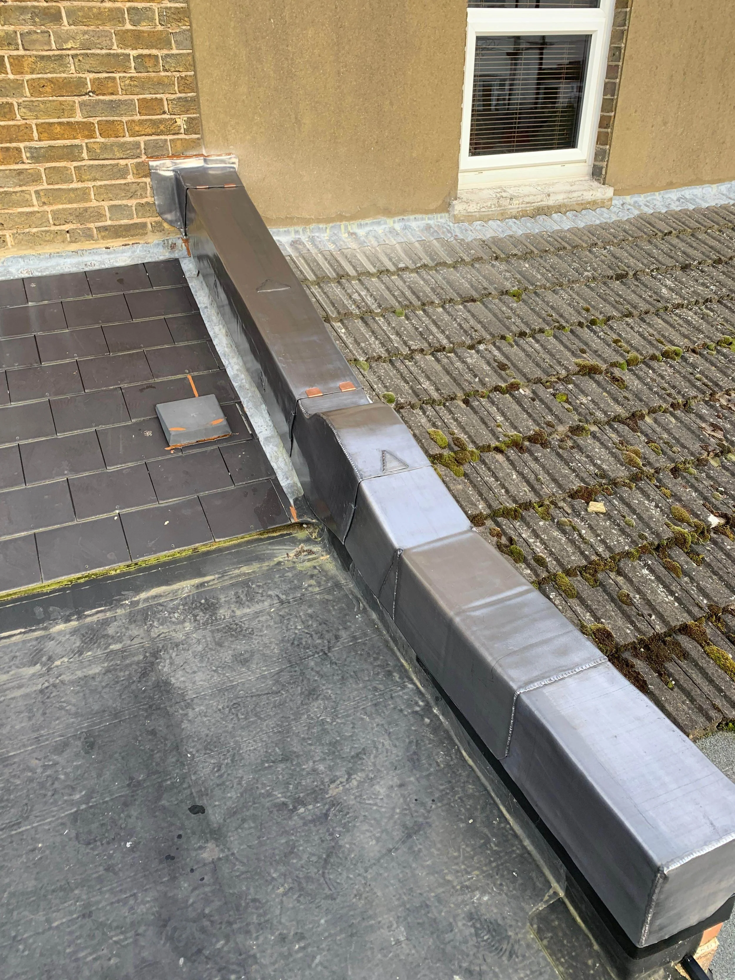 Roof with newly installed metal flashing and drainage system alongside older roof tiles, with a brick wall and a window in the background.