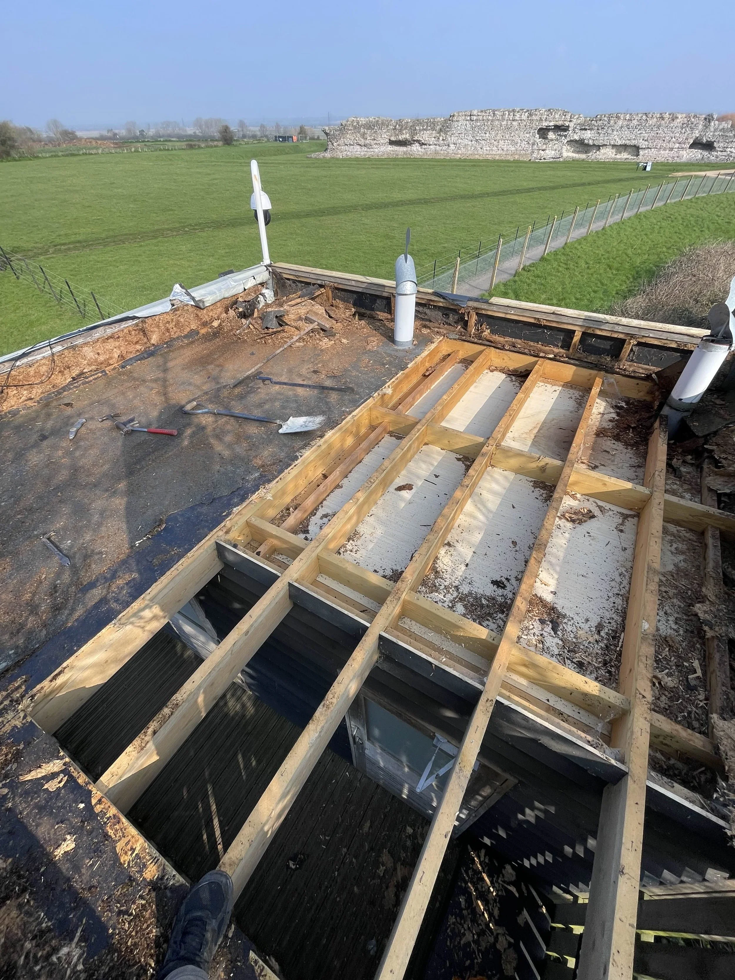 Rooftop under renovation showing wooden framing and attic space, with tools and open areas, overlooking a green field and distant stone ruins.