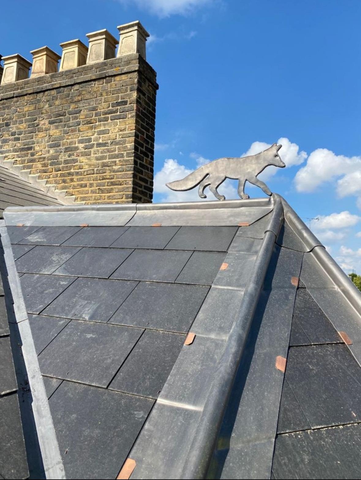 A rooftop with gray tiles and a metal fox sculpture standing on the ridge of the roof, with a brick chimney in the background and a blue sky with clouds.