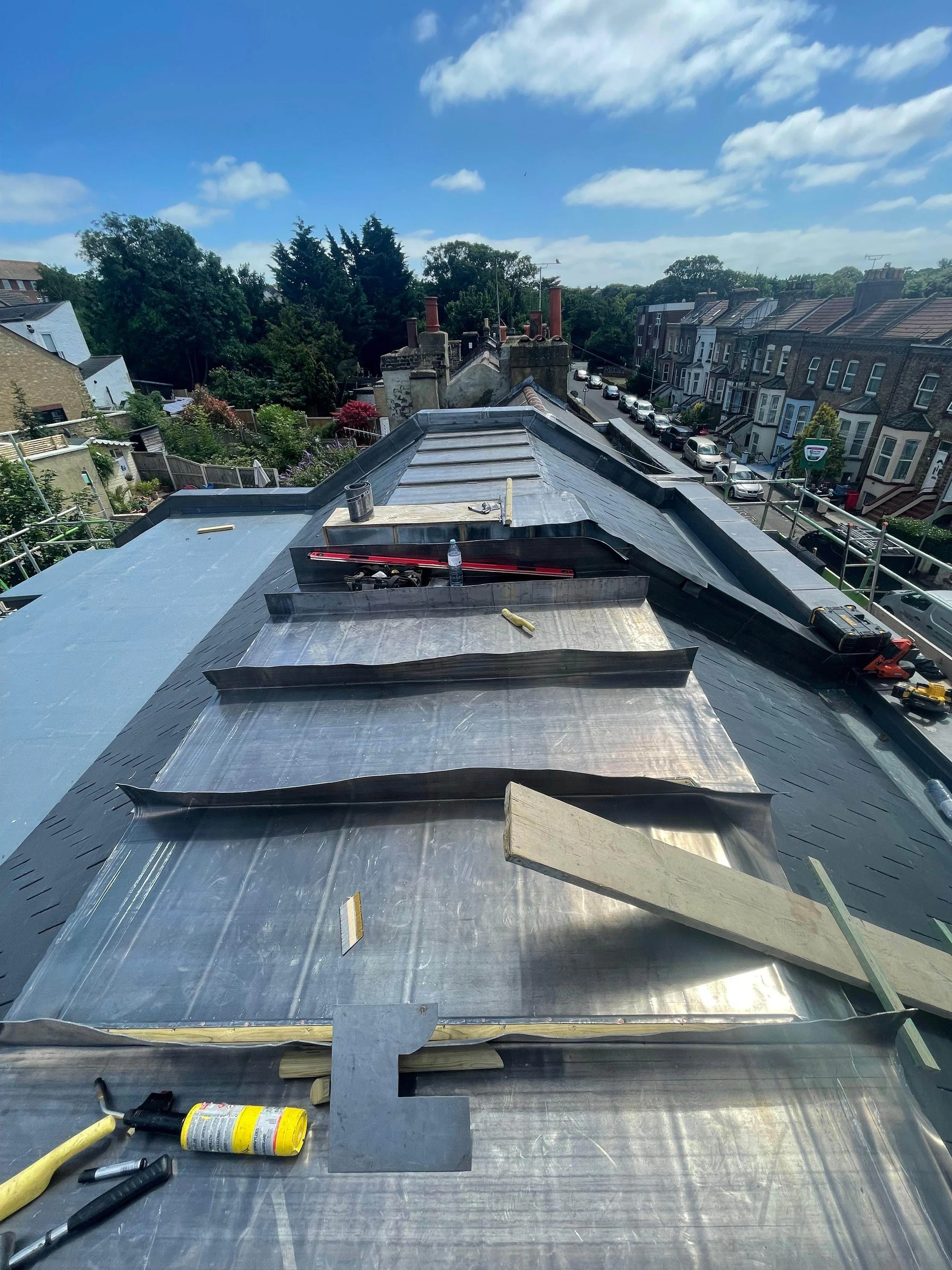 View of a roof under construction or repair, with metal panels and various tools on it, overlooking a neighborhood with residential buildings, trees, and a blue sky with clouds.