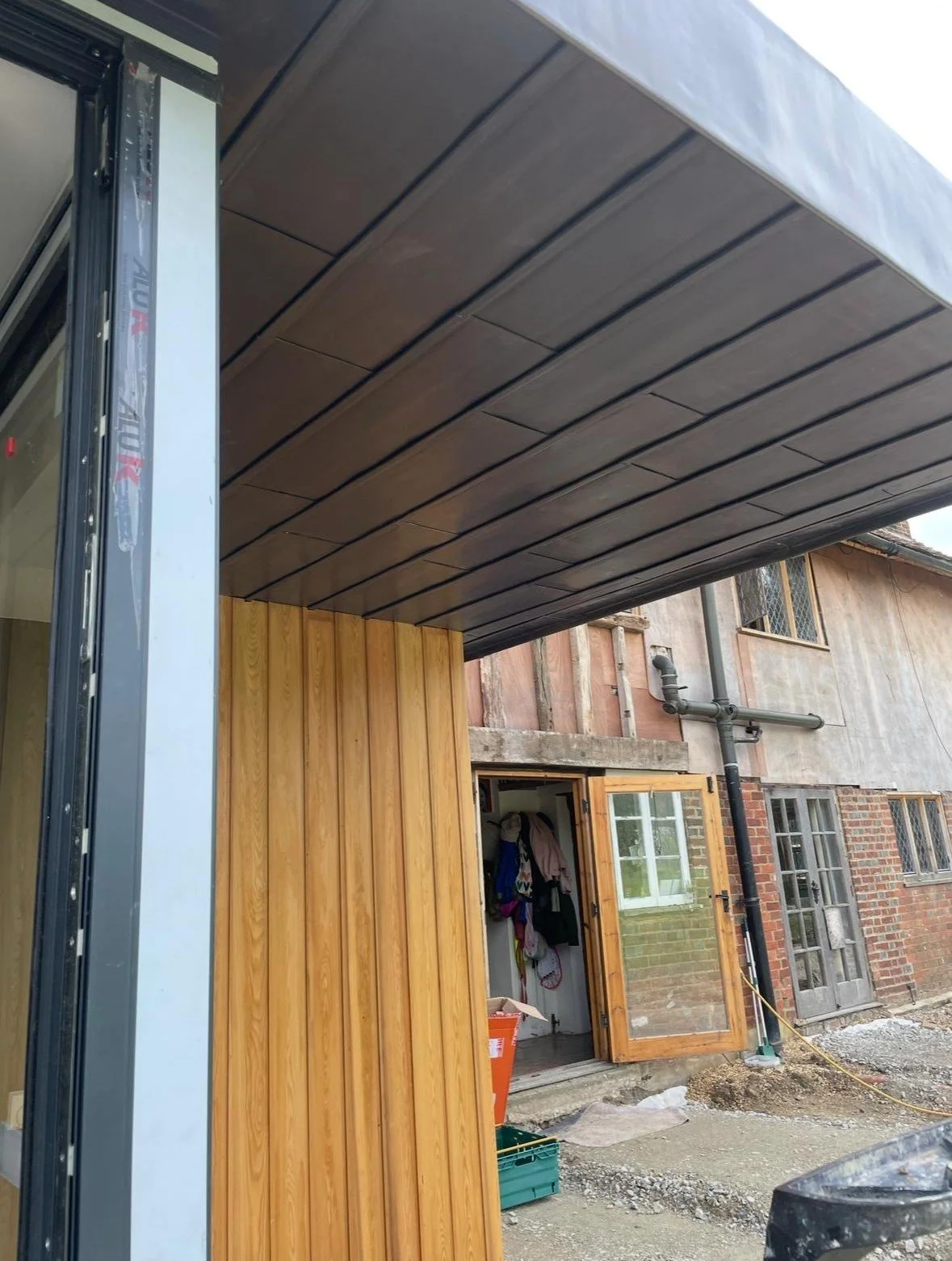 View of a house under construction with a wood and glass extension, showing a black roof overhanging a partly finished wooden wall and an open door revealing items inside.