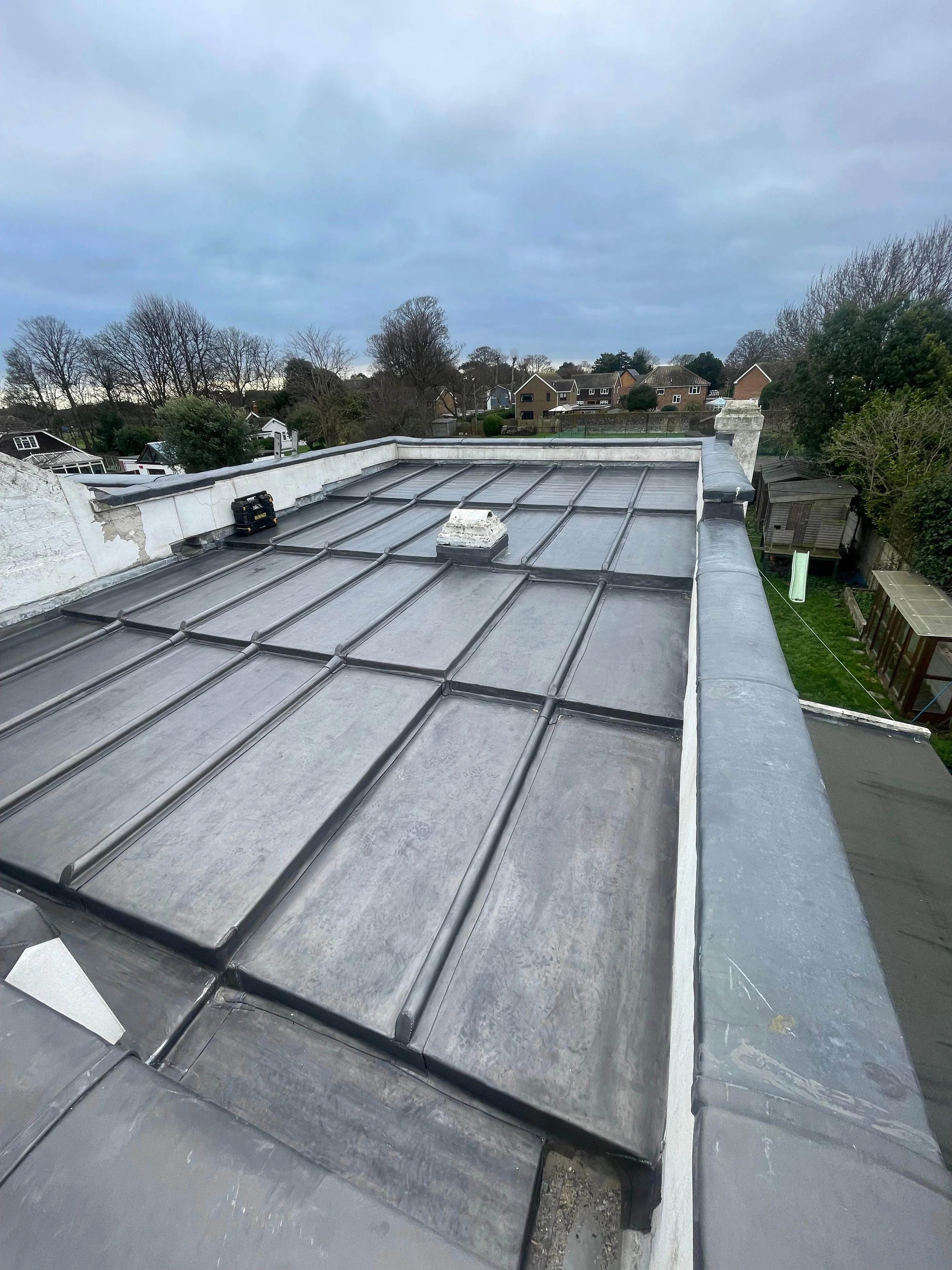 View of a flat metal roof with black coating, a small black toolbox, and white chimney on a residential building. Surrounding neighborhood with trees and houses under cloudy sky.