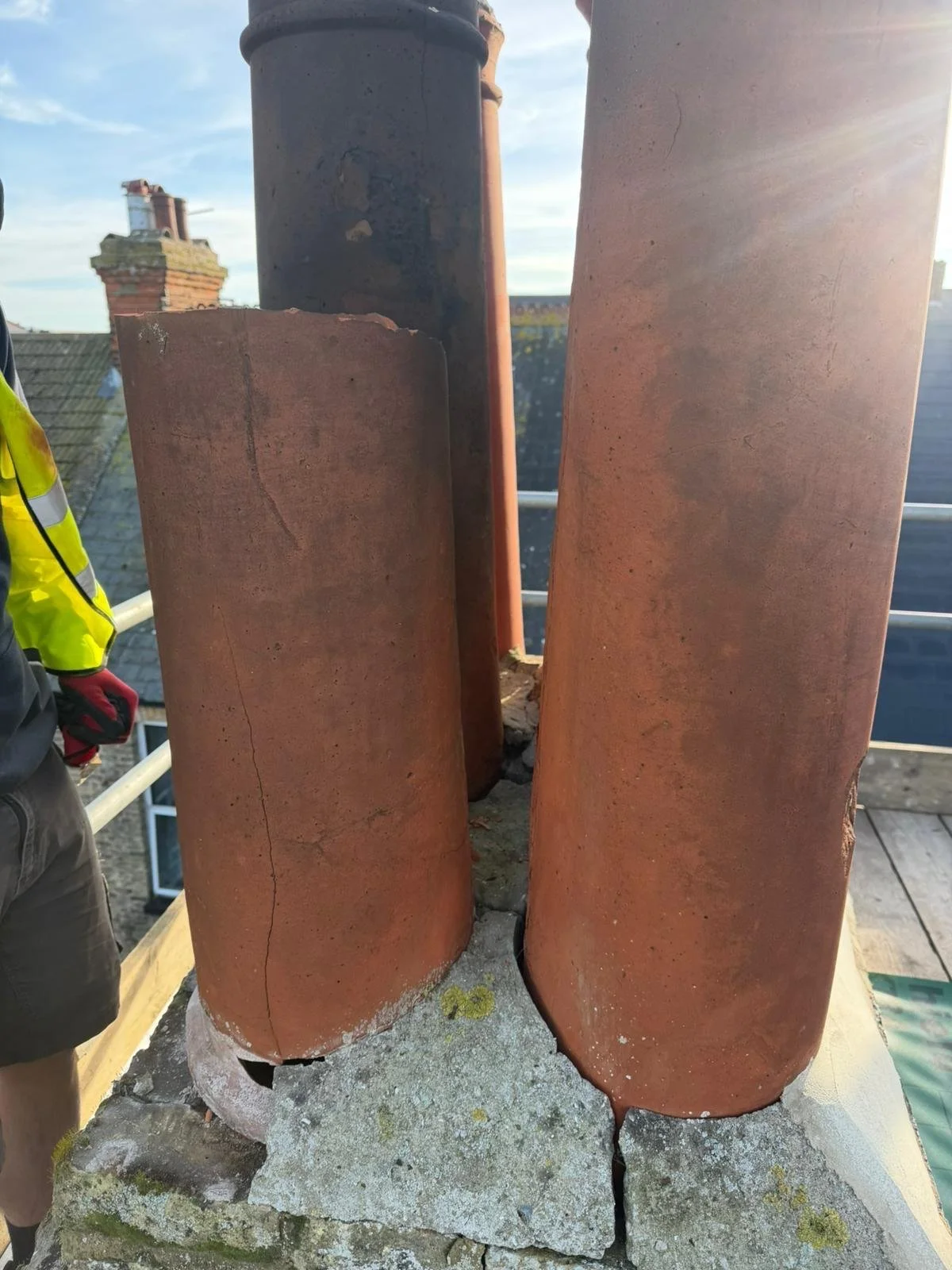 Close-up of three reddish-brown chimney flues on a rooftop with gray mortar and cracks, with part of a person's arm visible on the left.