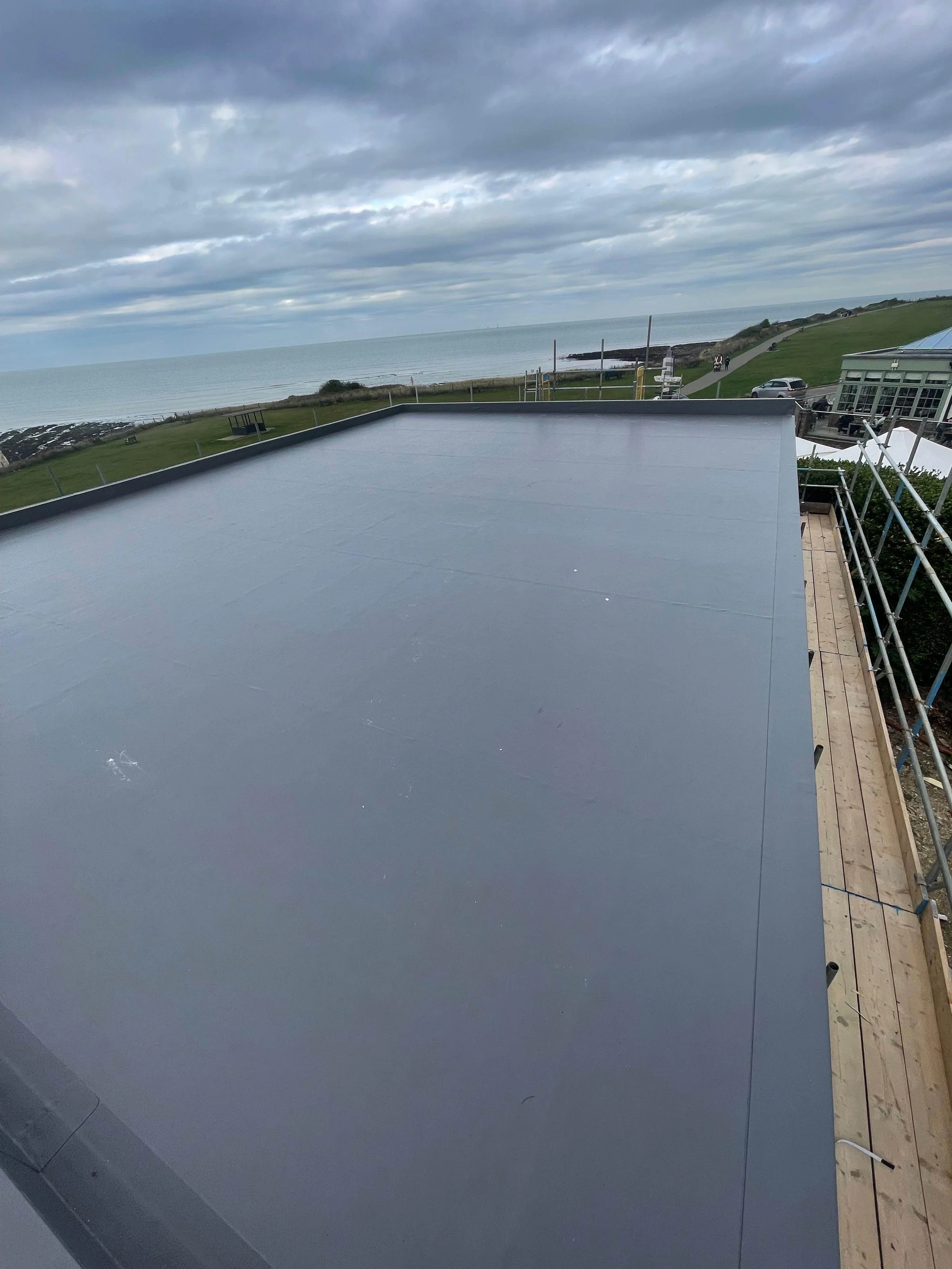 A flat rooftop with a gray surface overlooking a grassy area, ocean, and cloudy sky with a coastline in the distance.