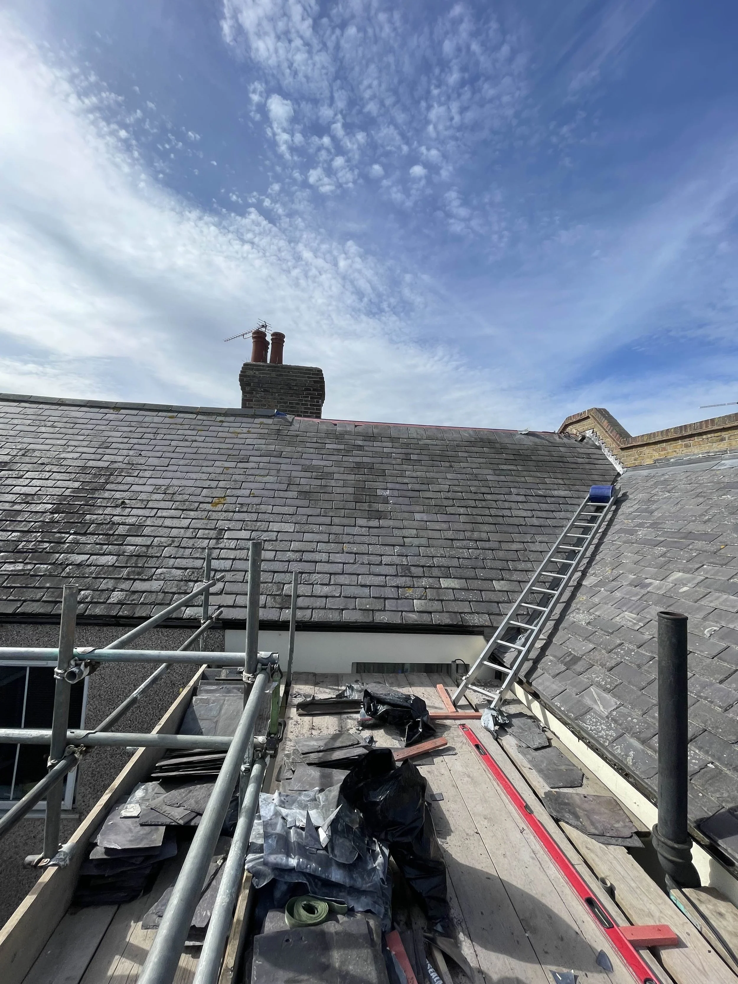 Construction work on a rooftop with tools, materials, and a ladder leading to the roof. The sky is partly cloudy.