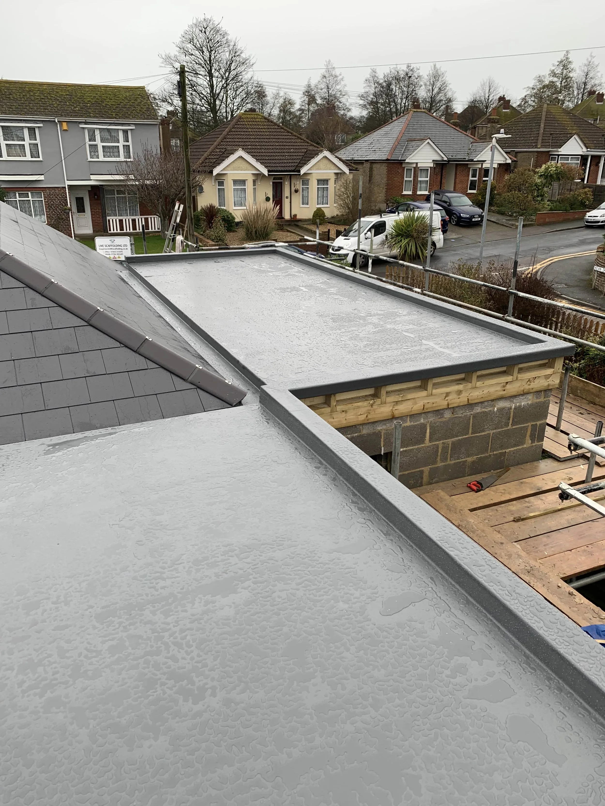 Rain-covered flat roofs under construction with residential houses and street visible in the background.