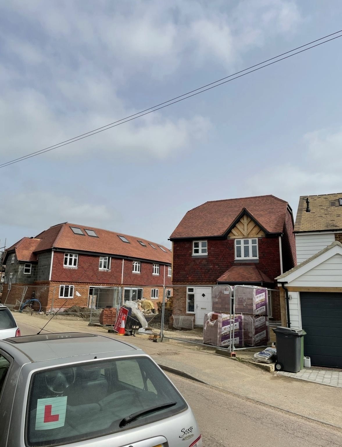 Residential street with construction work on houses, cars parked on the roadside, and a partly cloudy sky.