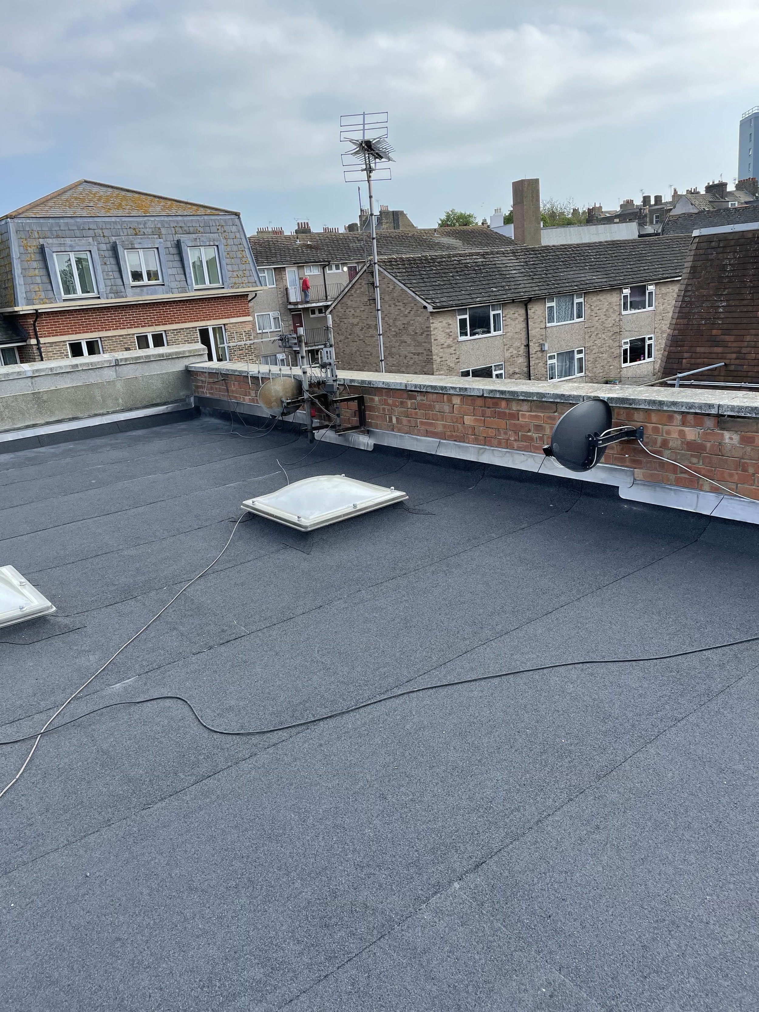 Rooftop with antennas, satellite dishes, and skylights, overlooking residential buildings and a cloudy sky.