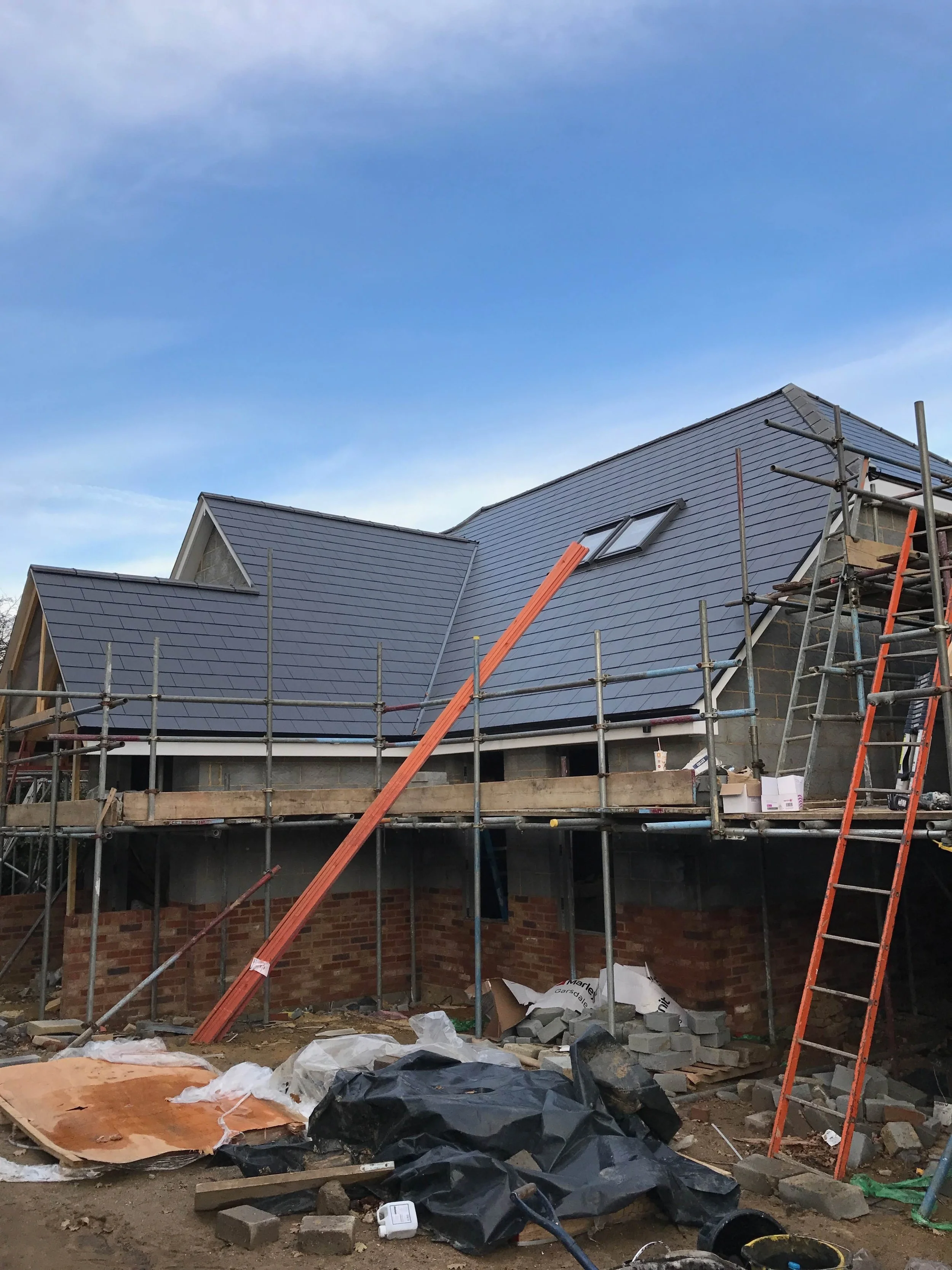 Construction site with a house under renovation, scaffolding around the building, finished roof, and construction materials and equipment on the ground.