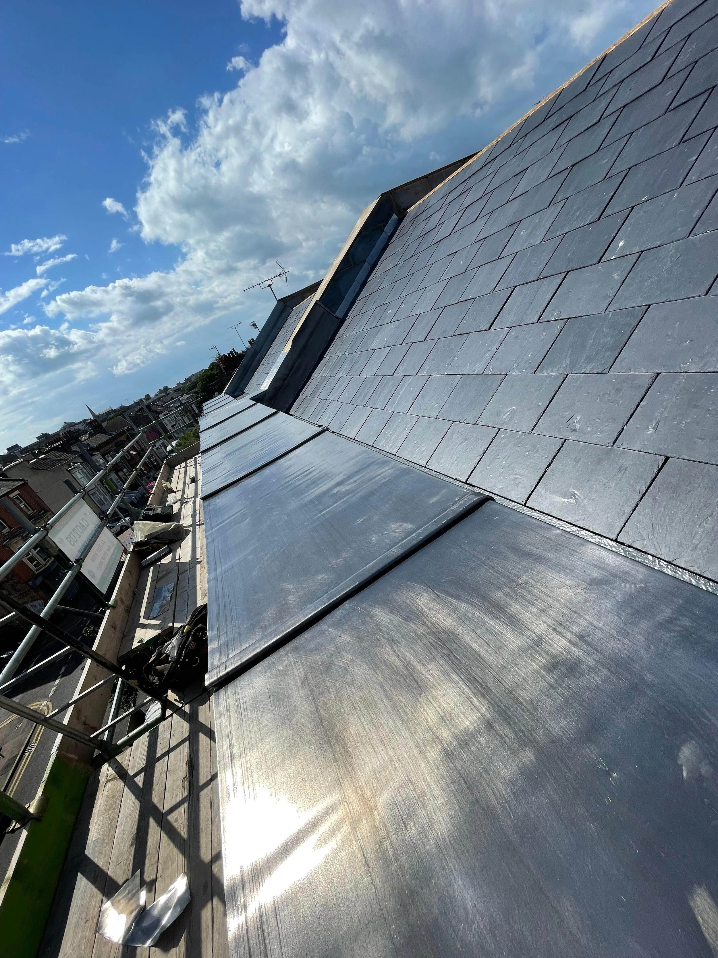 Photograph of a sloped roof with slate tiles and metal flashing, under a partly cloudy blue sky.