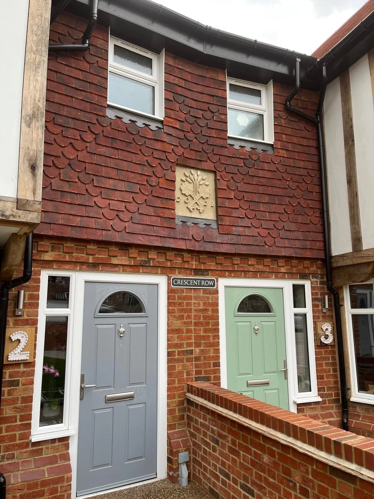 A brick residential building with two front doors, one blue and one green, on Crescent Row street. Above the doors, there are two small windows, and a decorative sign with a tree and the year 2021. The building has black drainage pipes and wooden acc