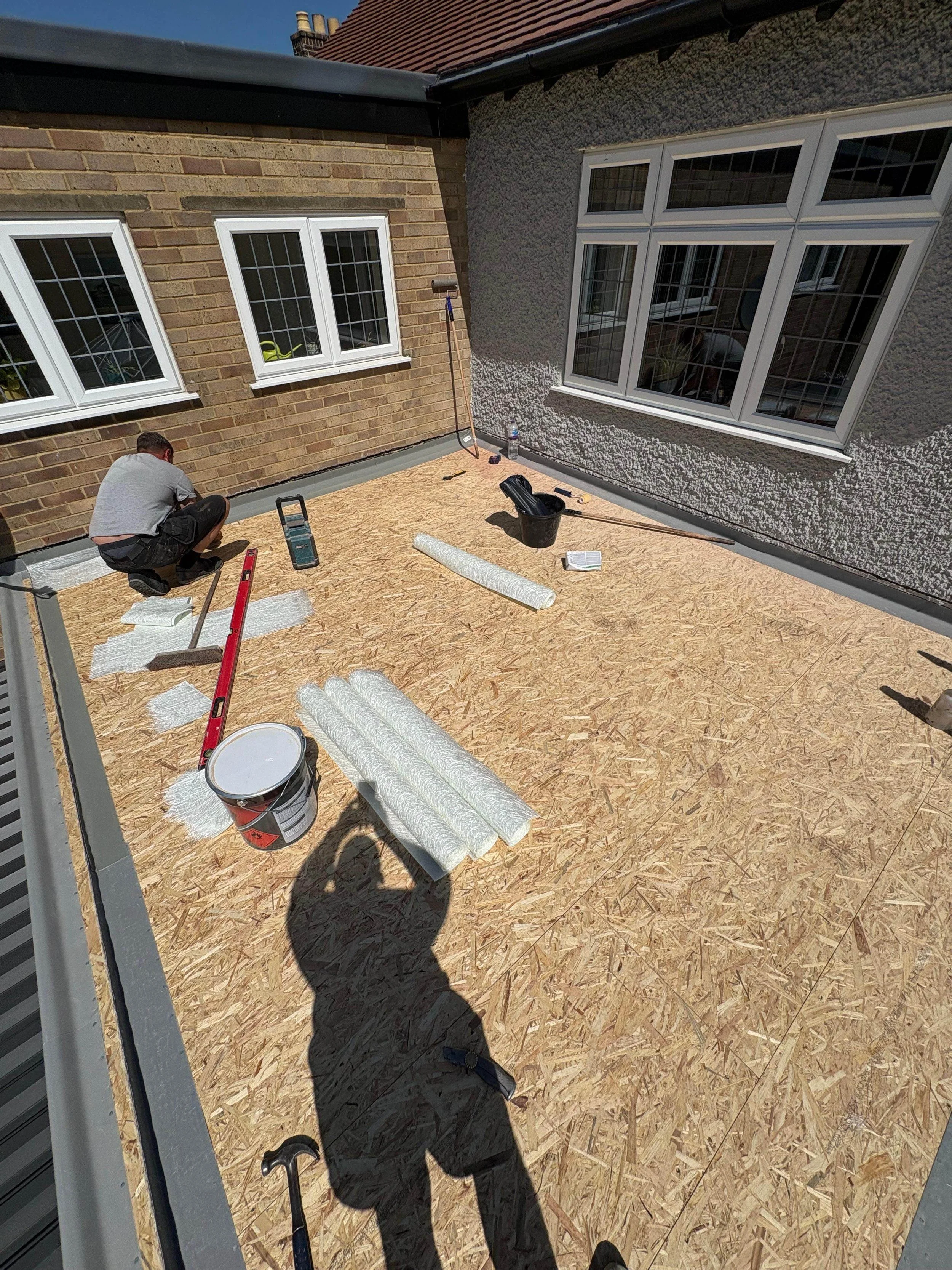 Construction worker installing or repairing a roof with plywood sheathing, surrounded by tools and materials, on a sunny day.