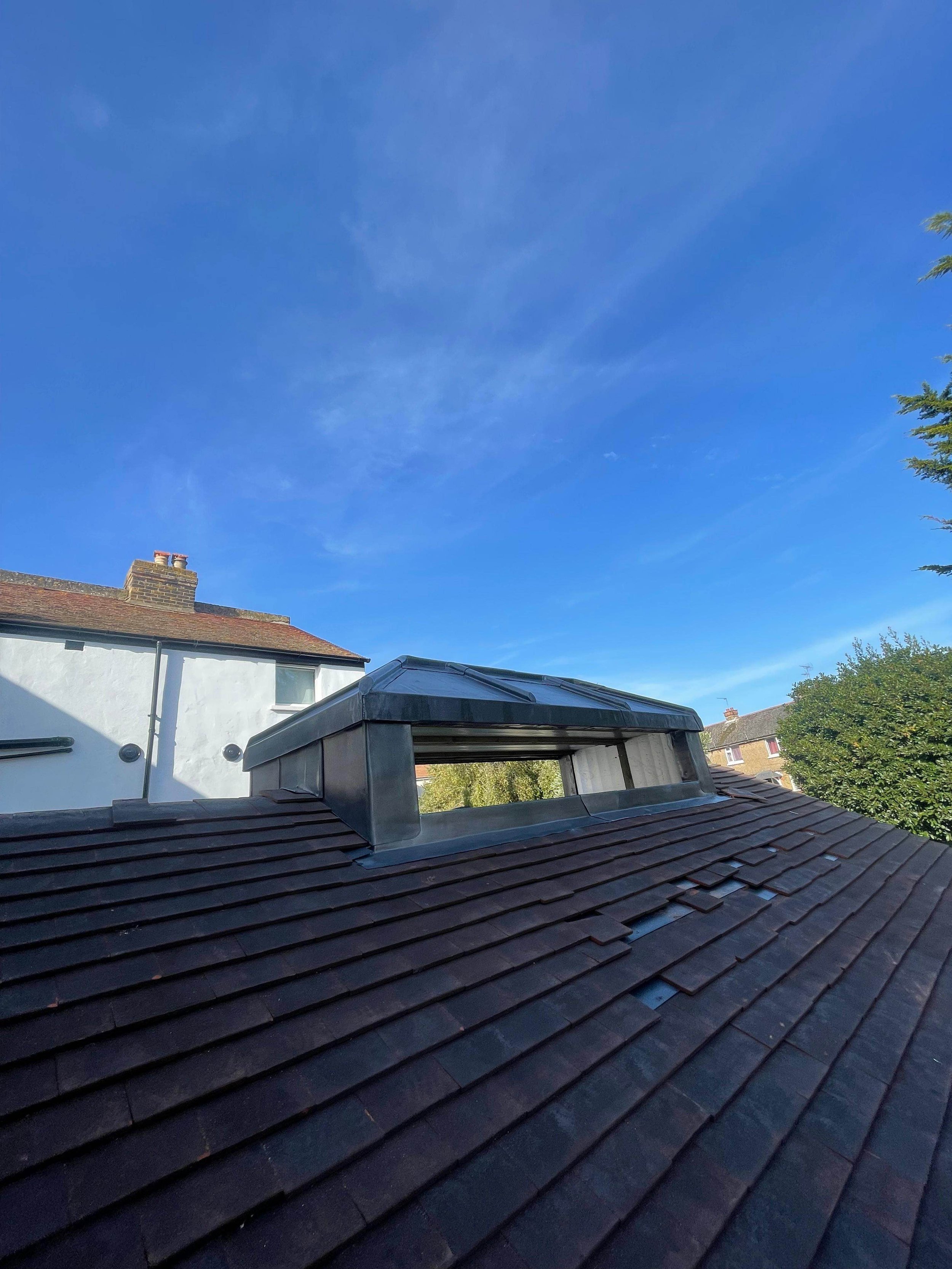 View of a roof with a dormer window under a blue sky with a few clouds, neighboring houses, and trees in the background.