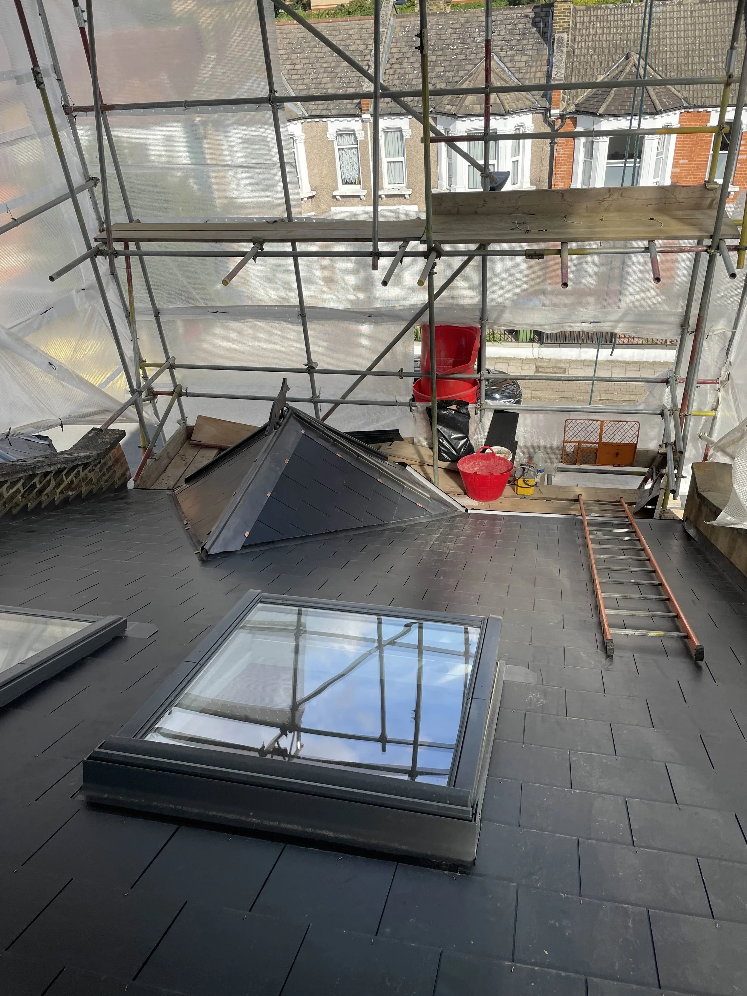 View from a rooftop under renovation featuring black tiled roof, retrofitted skylight window, scaffolding, and various construction tools and materials.