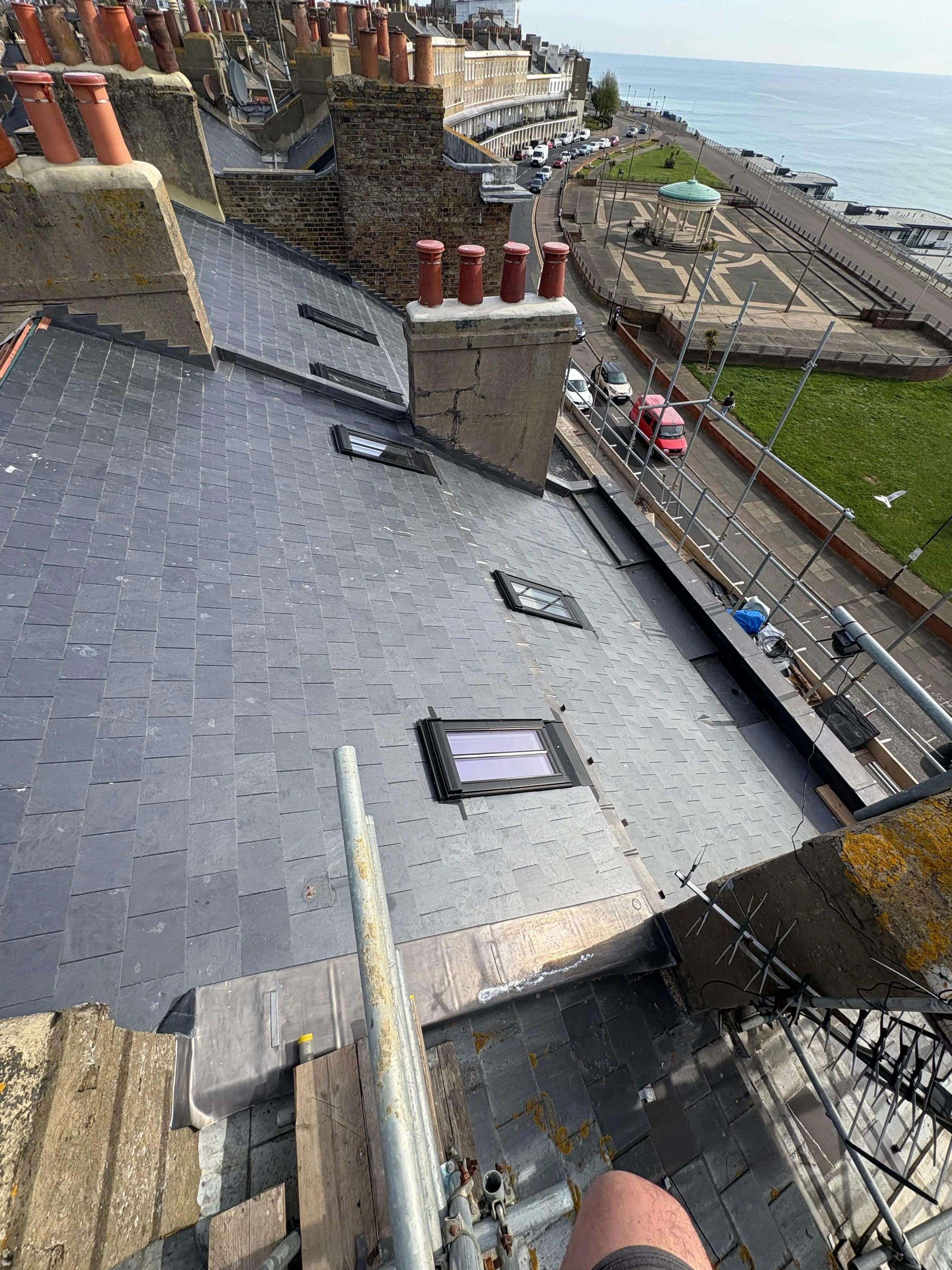 View of a rooftop with four skylights, surrounding chimneys, and construction scaffolding, overlooking a street with parked cars, a grassy park with a gazebo, and the ocean in the background.