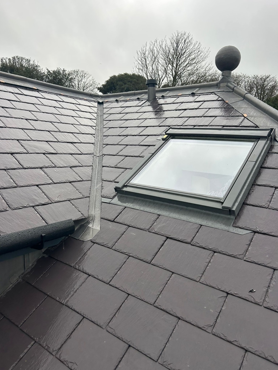 Close-up of a rooftop with slate tiles, showing a skylight window and a vent pipe on a cloudy day.