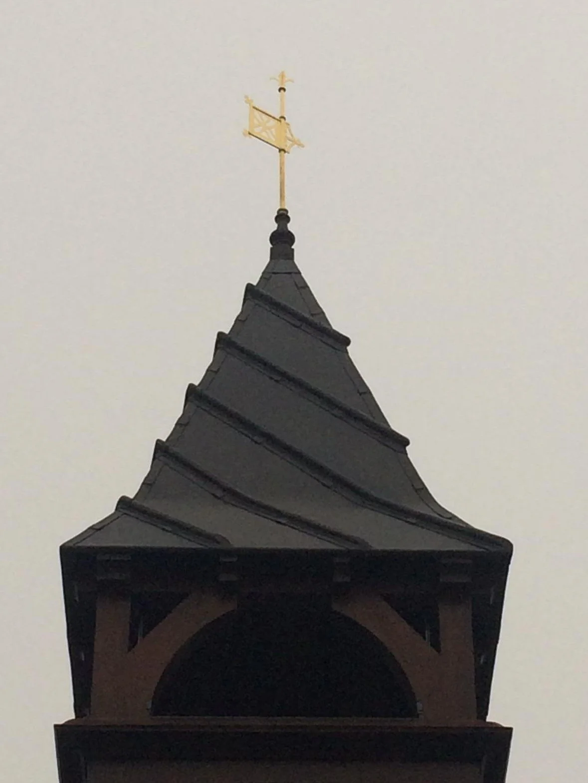 Close-up of a church steeple with a gold weather vane on top, set against a gray sky.