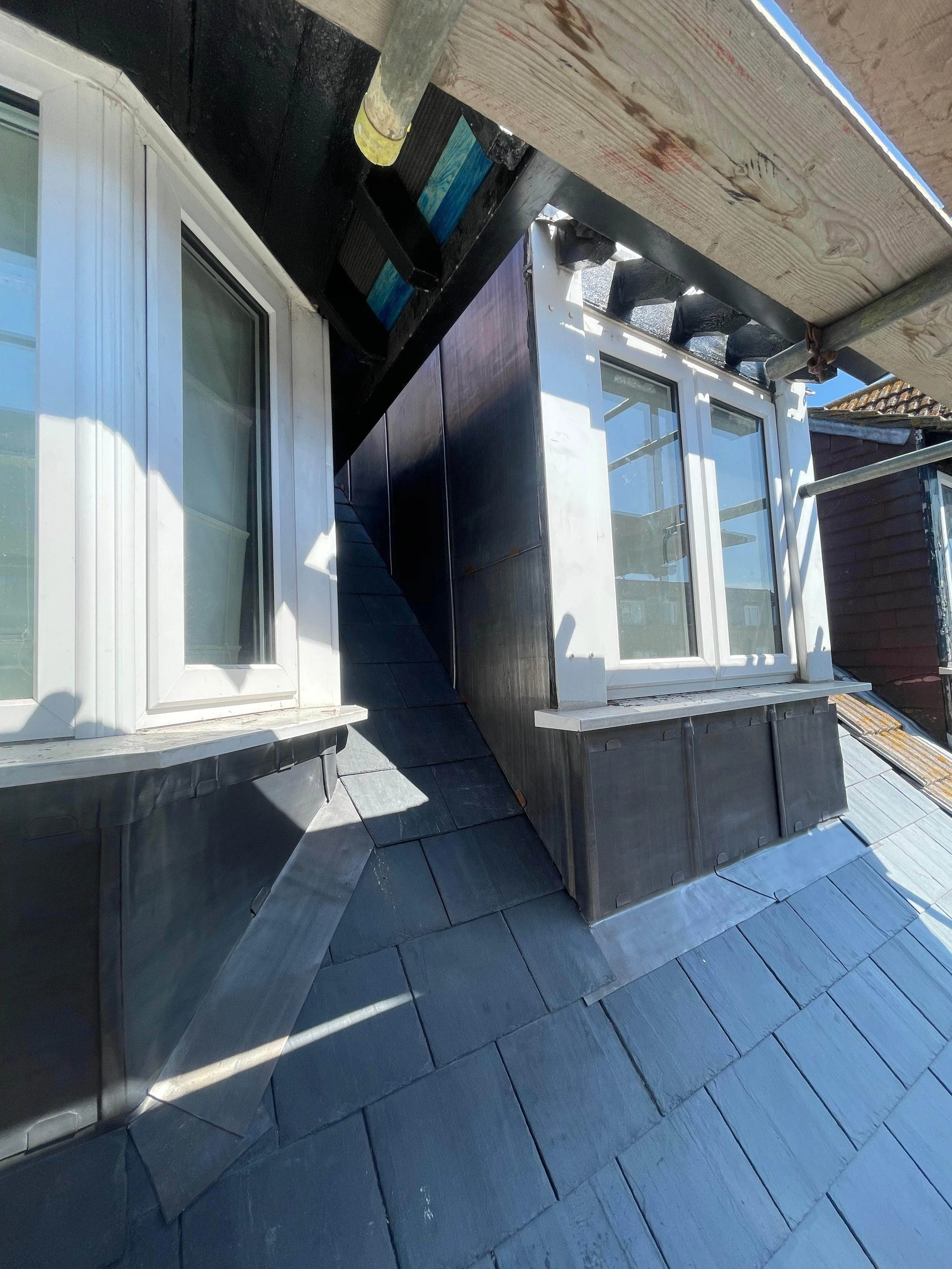 View of a rooftop with two white-framed windows, black siding, and slate shingles. Construction scaffolding and wood beams are visible.