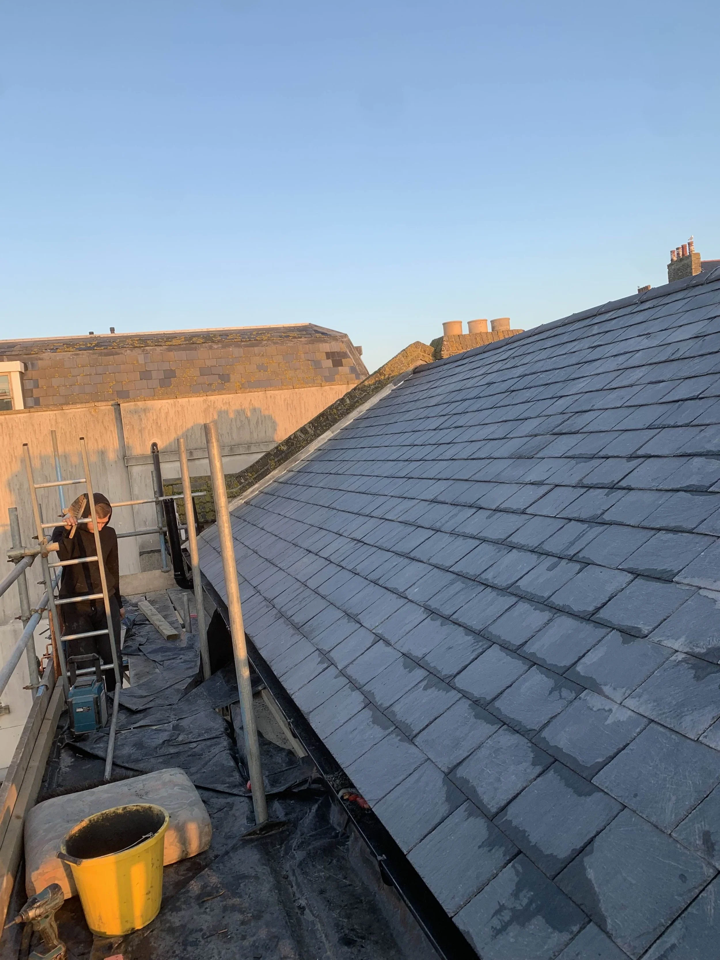 A worker standing on a scaffold next to a house roof, installing or repairing slate shingles during daylight.