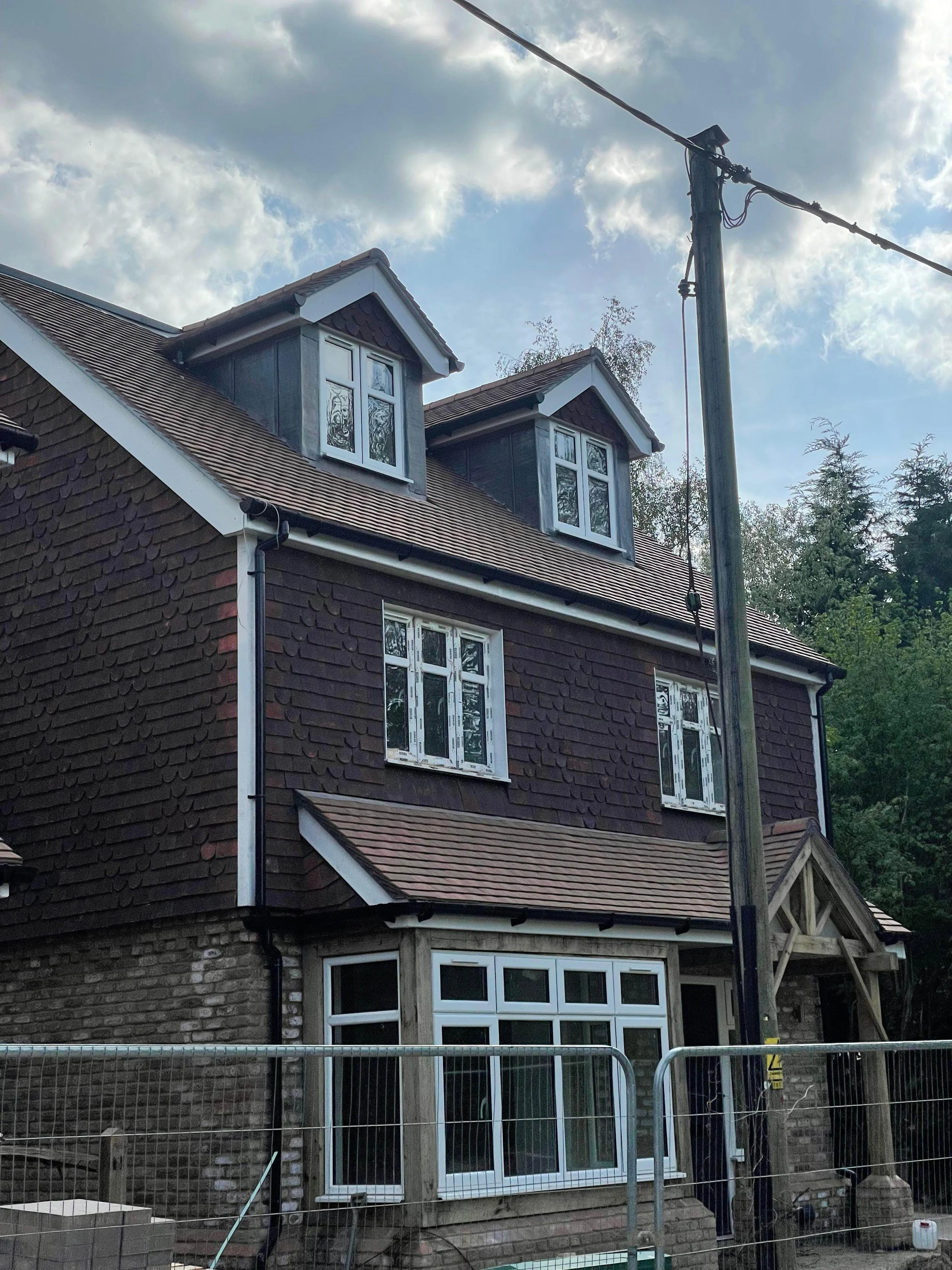 A multi-story house under construction with dark brown shingles, white window frames, and a front porch; a construction barrier surrounds the site; a power pole with wires stands nearby, and clouds are visible in the sky.