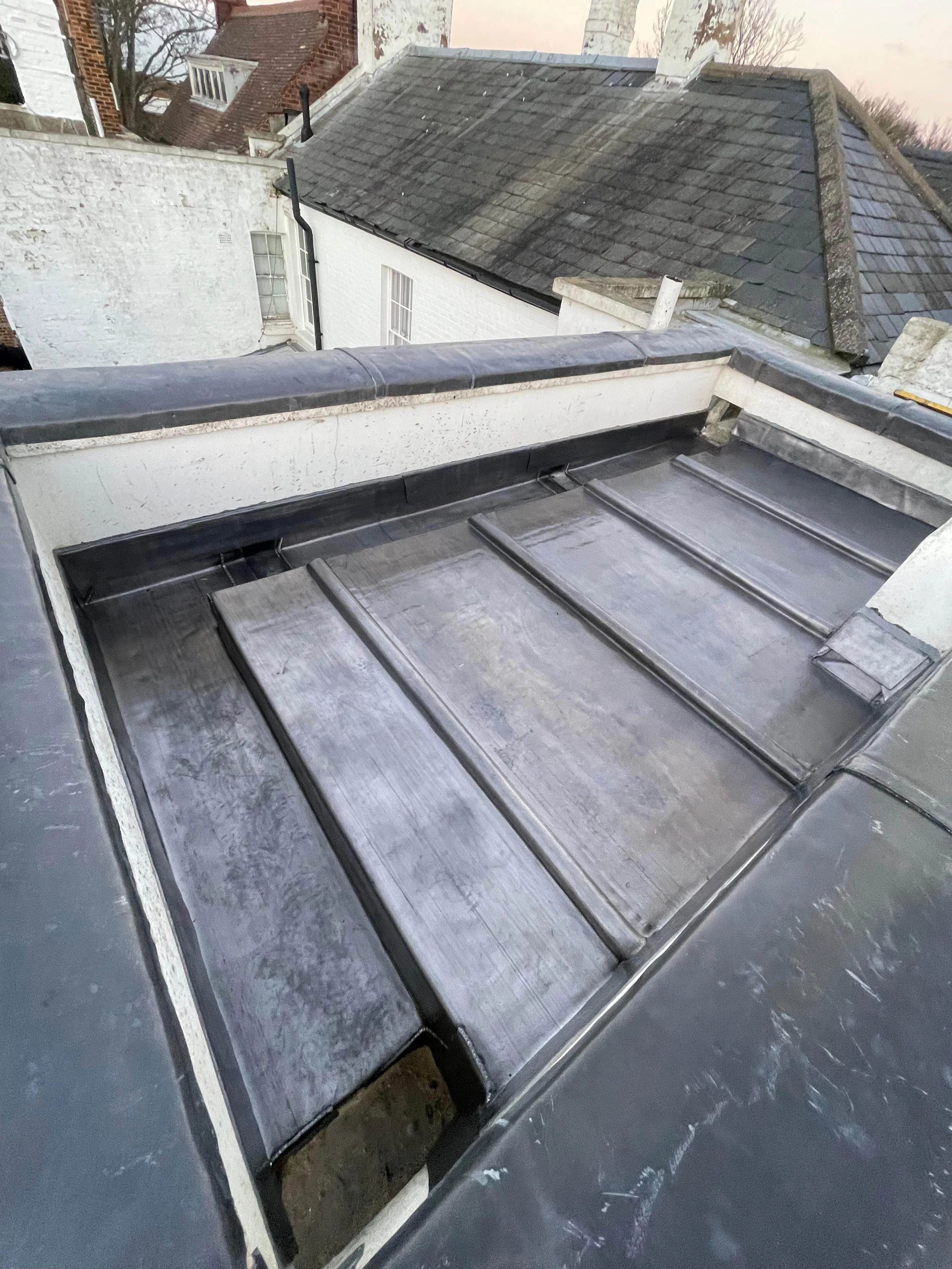 Metal roof with raised seams and neighboring rooftops and chimney flues in the background.