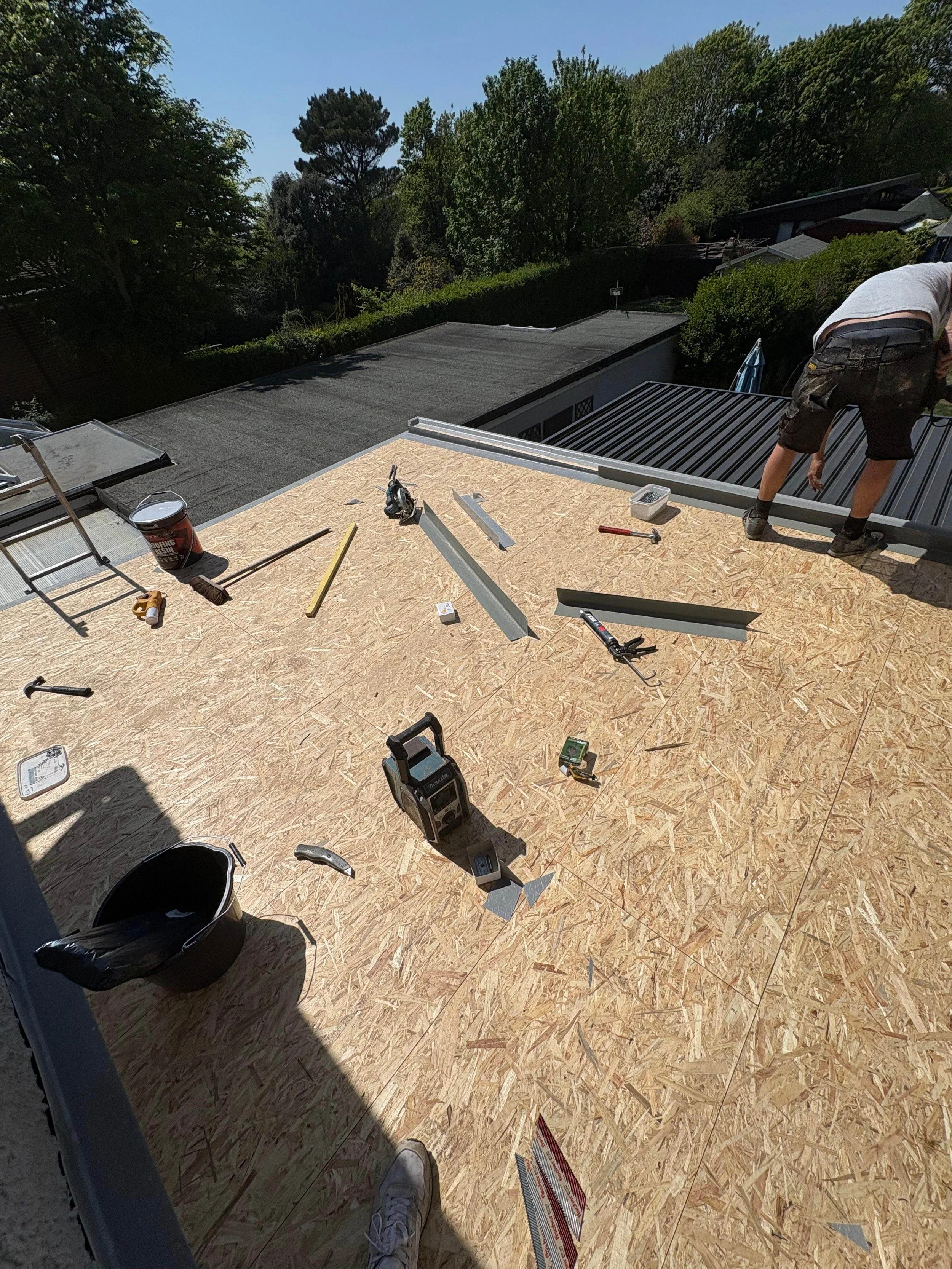 Construction worker installing a roof with oriented strand board (OSB) panels, various tools and materials scattered on the roof surface, and green trees in the background.