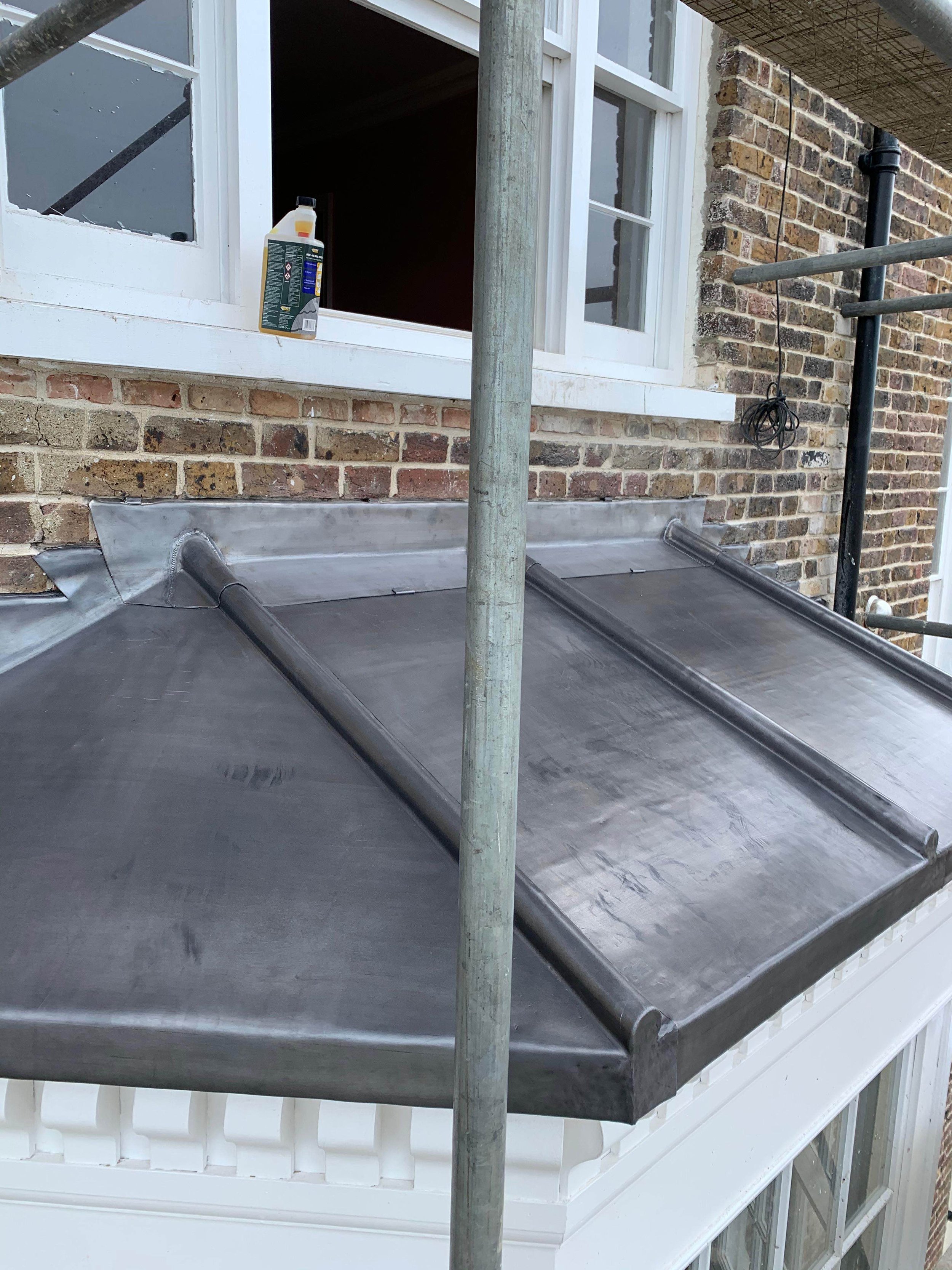 Roofing under construction with a black waterproof membrane, scaffolding poles, and an open window with a cleaning product on the sill.