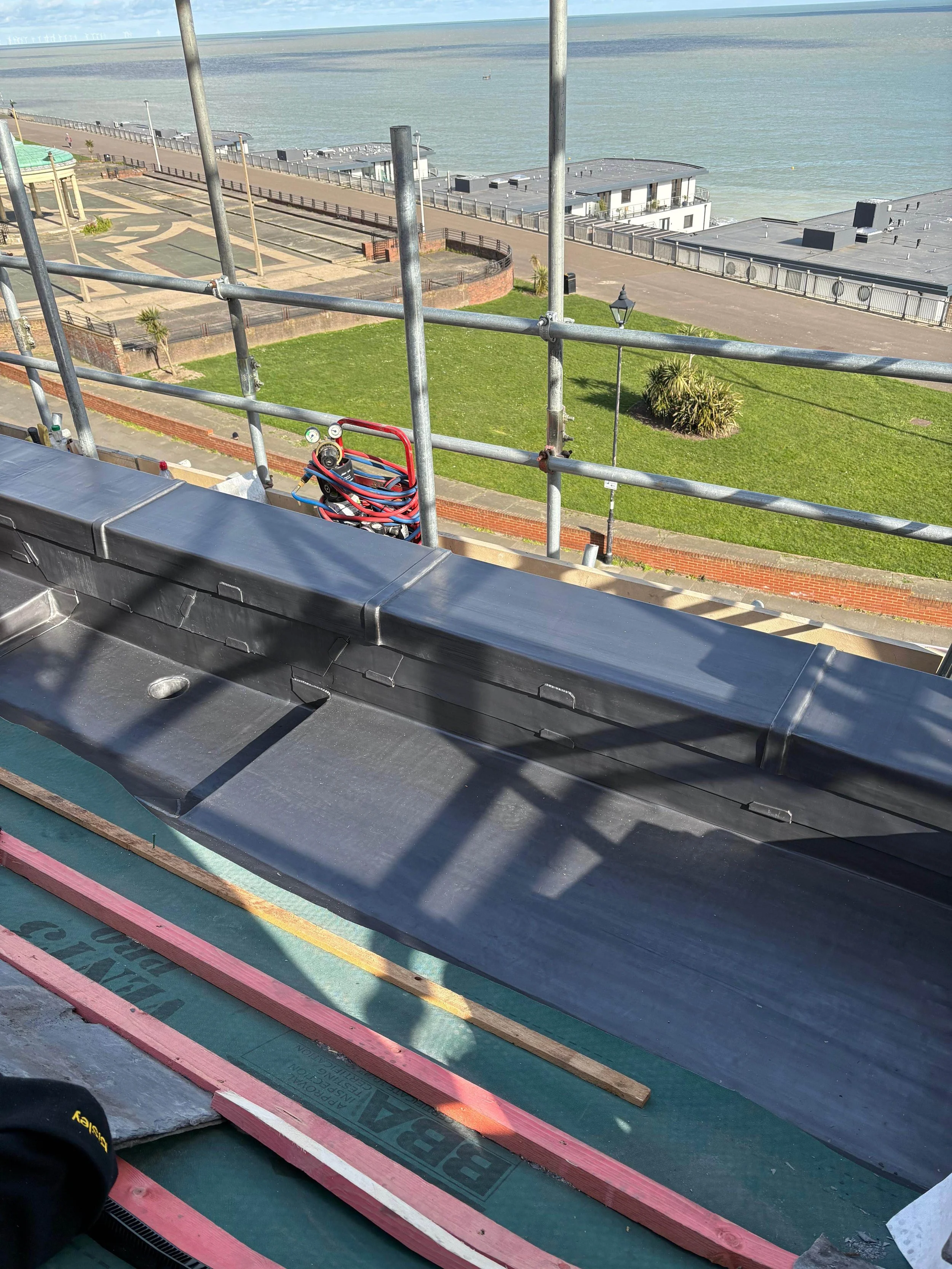 View from a building under construction showing scaffolding, roofing materials, and a coastal scene with grassy park, water, and buildings.