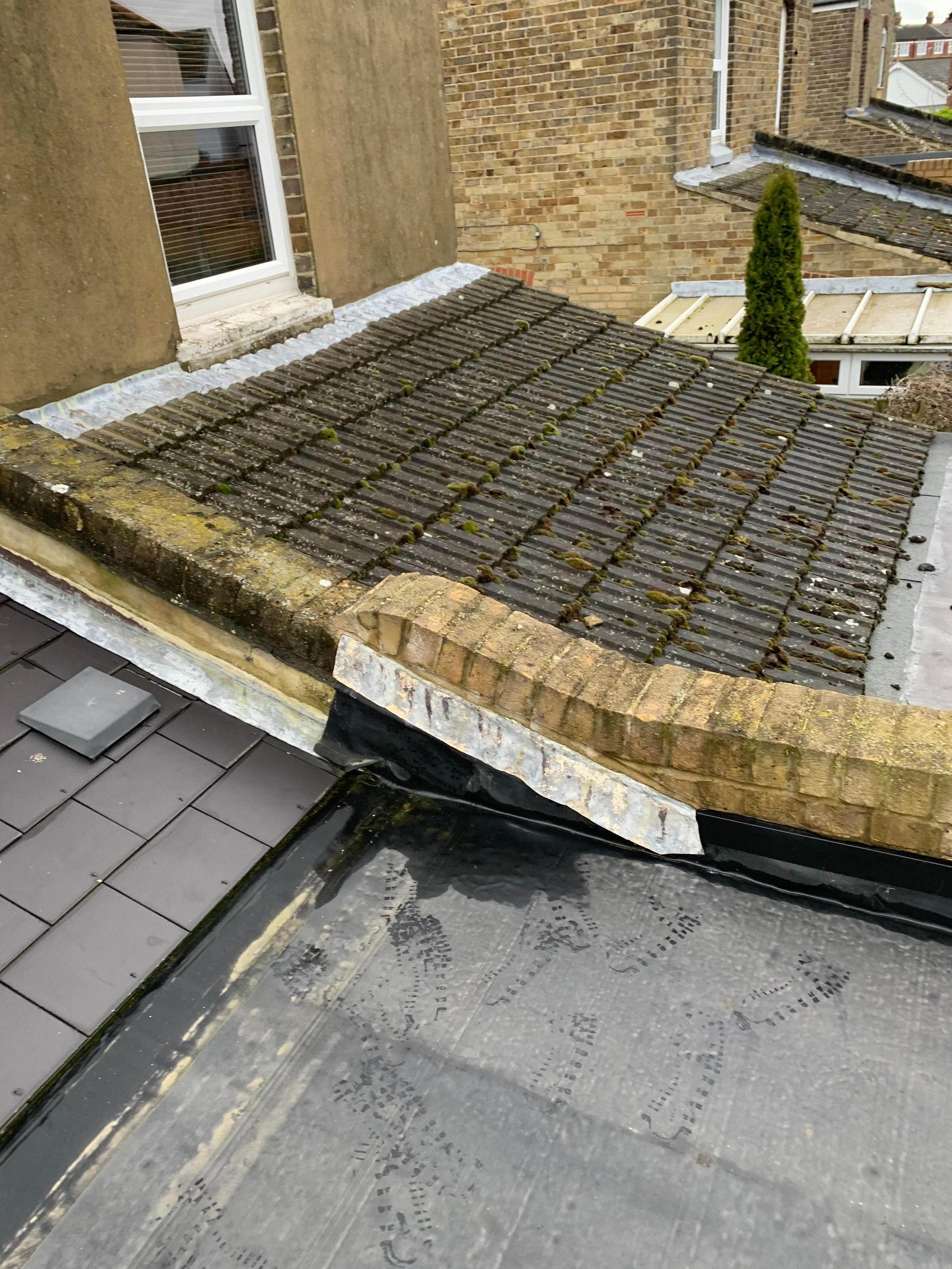 View of three different types of roofs on neighboring buildings, including a moss-covered tiled roof, a flat roof with black waterproofing material, and a small section of glass roof with metal framing, all in an urban area with brick walls and a win