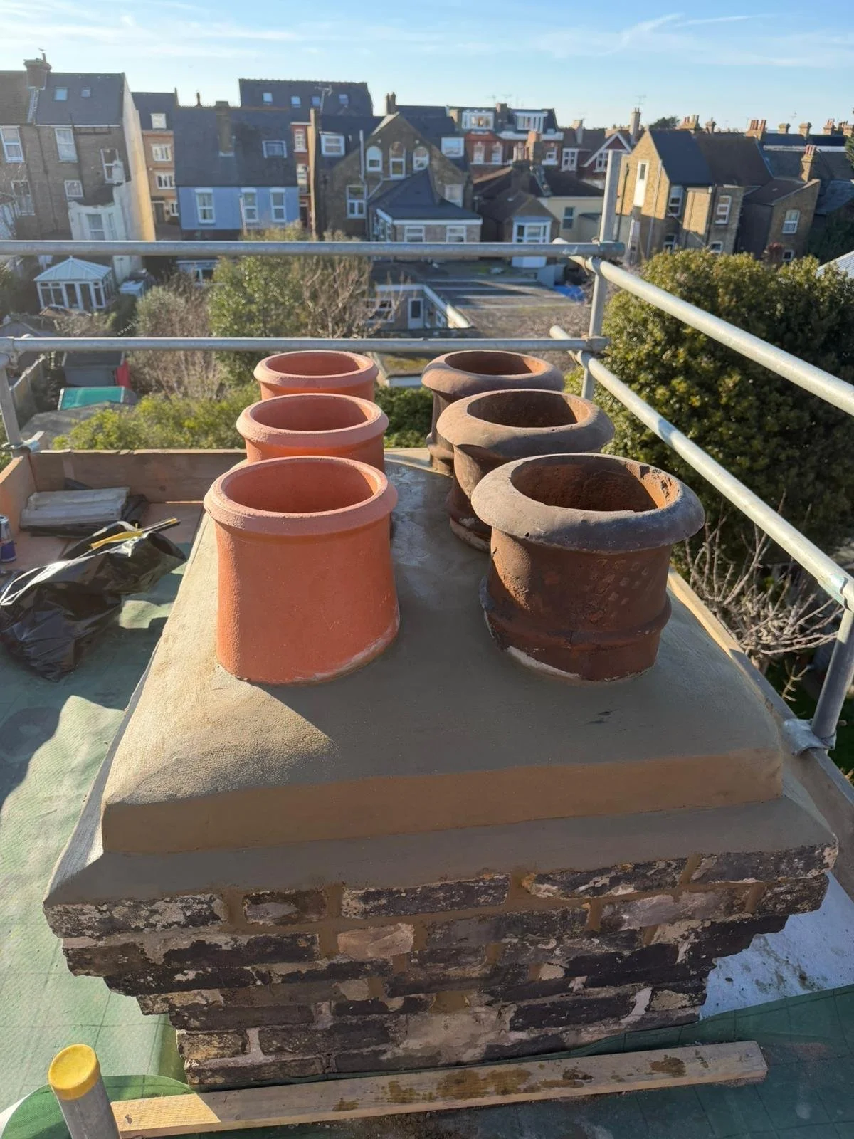 Six clay chimney pots on a brick chimney, with a view of a residential neighborhood with houses and trees in the background.