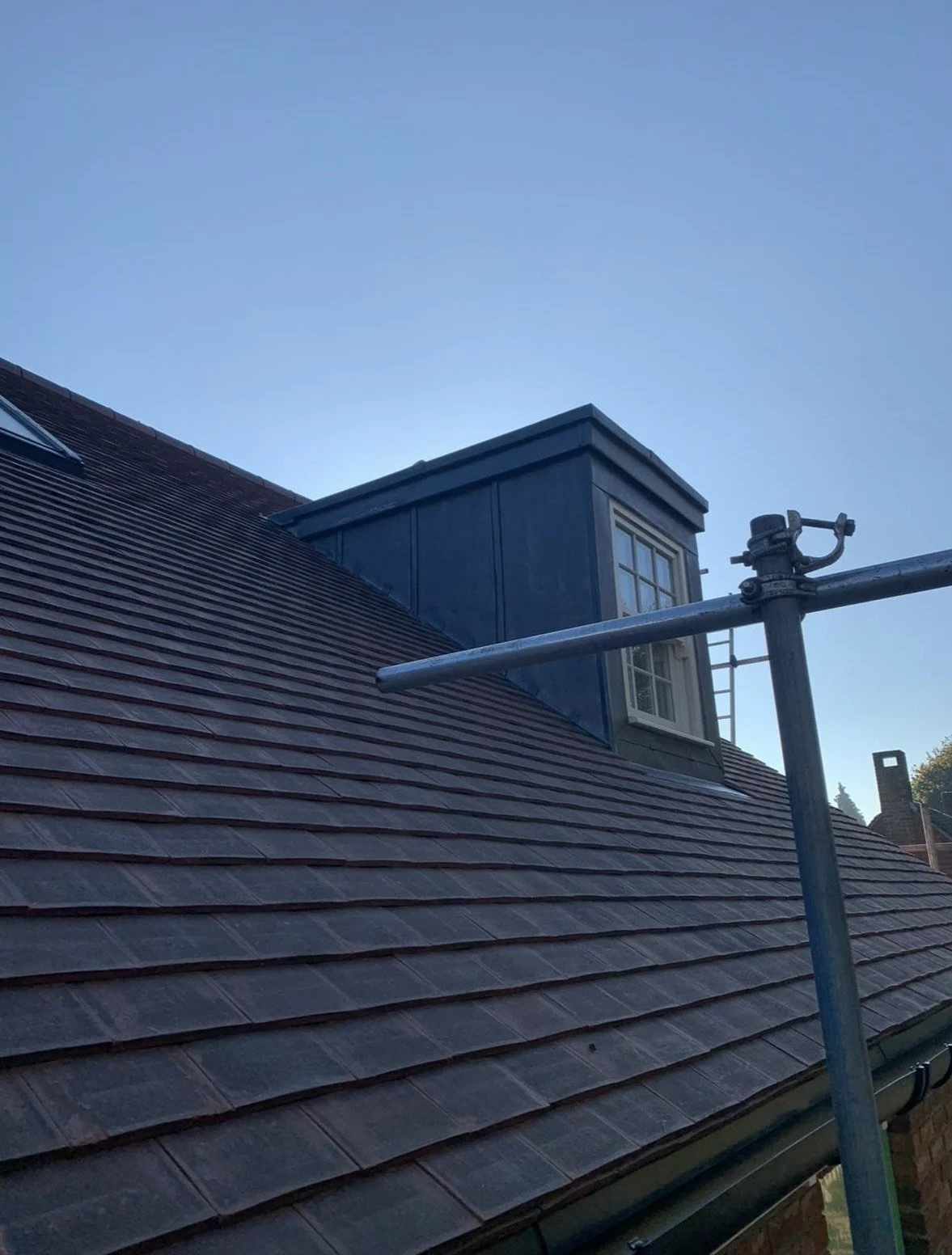 View of a sloped roof with reddish-brown tiles, a small blue dormer with a window, and scaffolding in the foreground, under a clear blue sky.