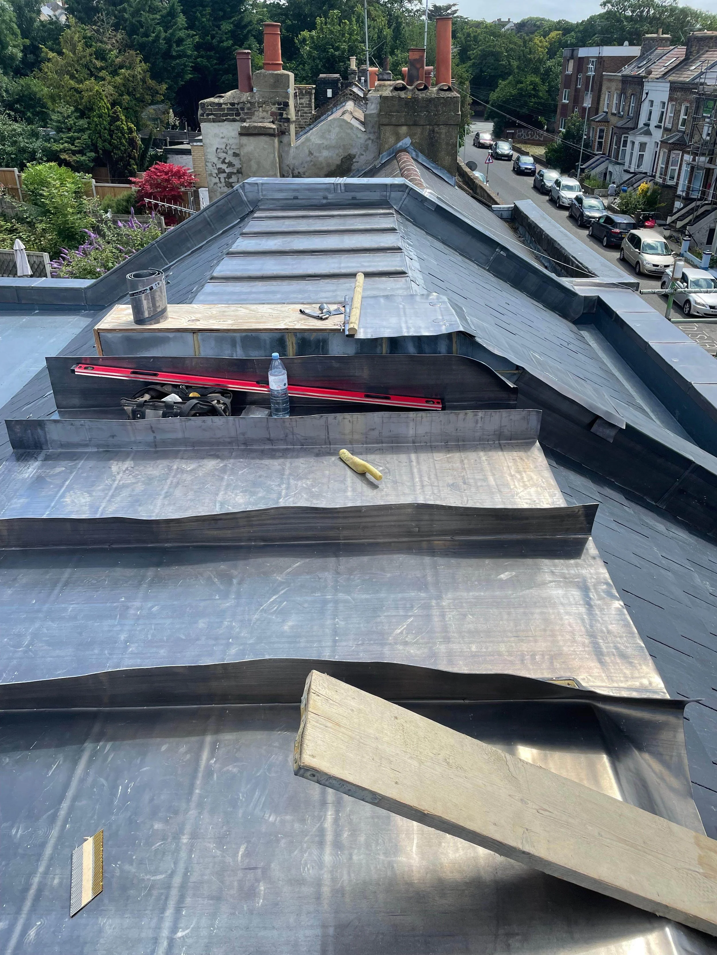 View of a metal roof under repair with tools, a level, water bottle, and wood plank on top, in a residential neighborhood with trees and parked cars.