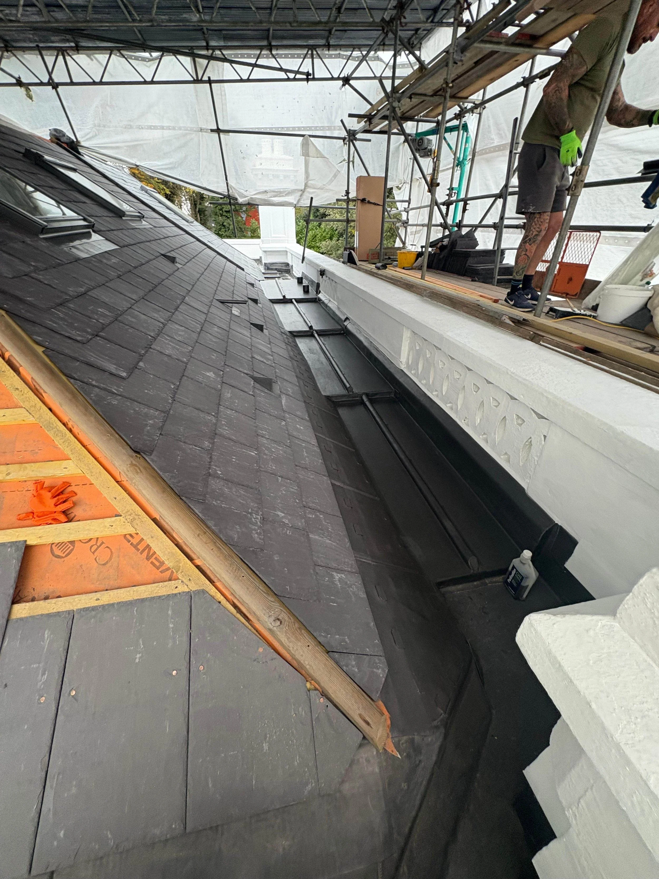 Construction workers on scaffolding repairing a sloped roof with black shingles and a white decorative wall underneath.