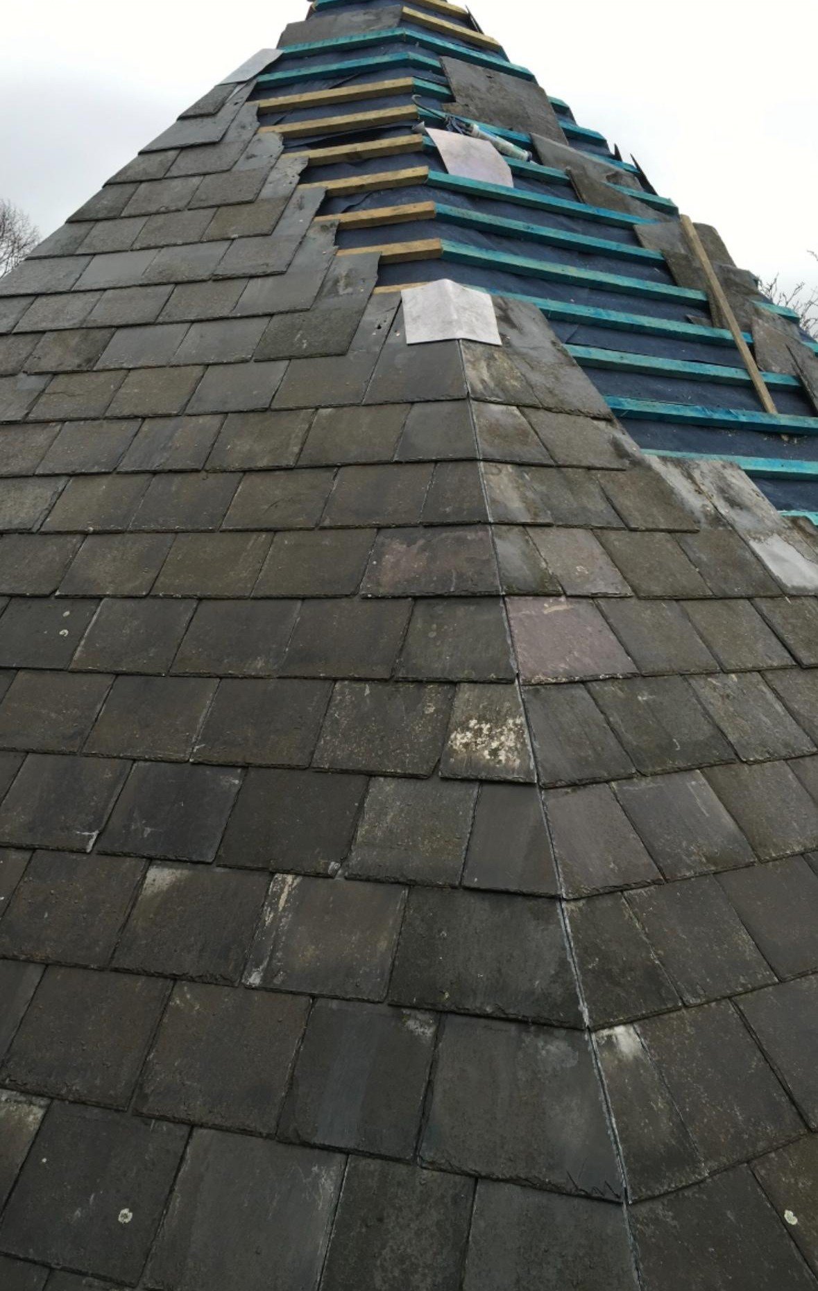 A roof under construction with layered shingles, some sections with added wooden battens and partially installed blue weatherproofing membrane.