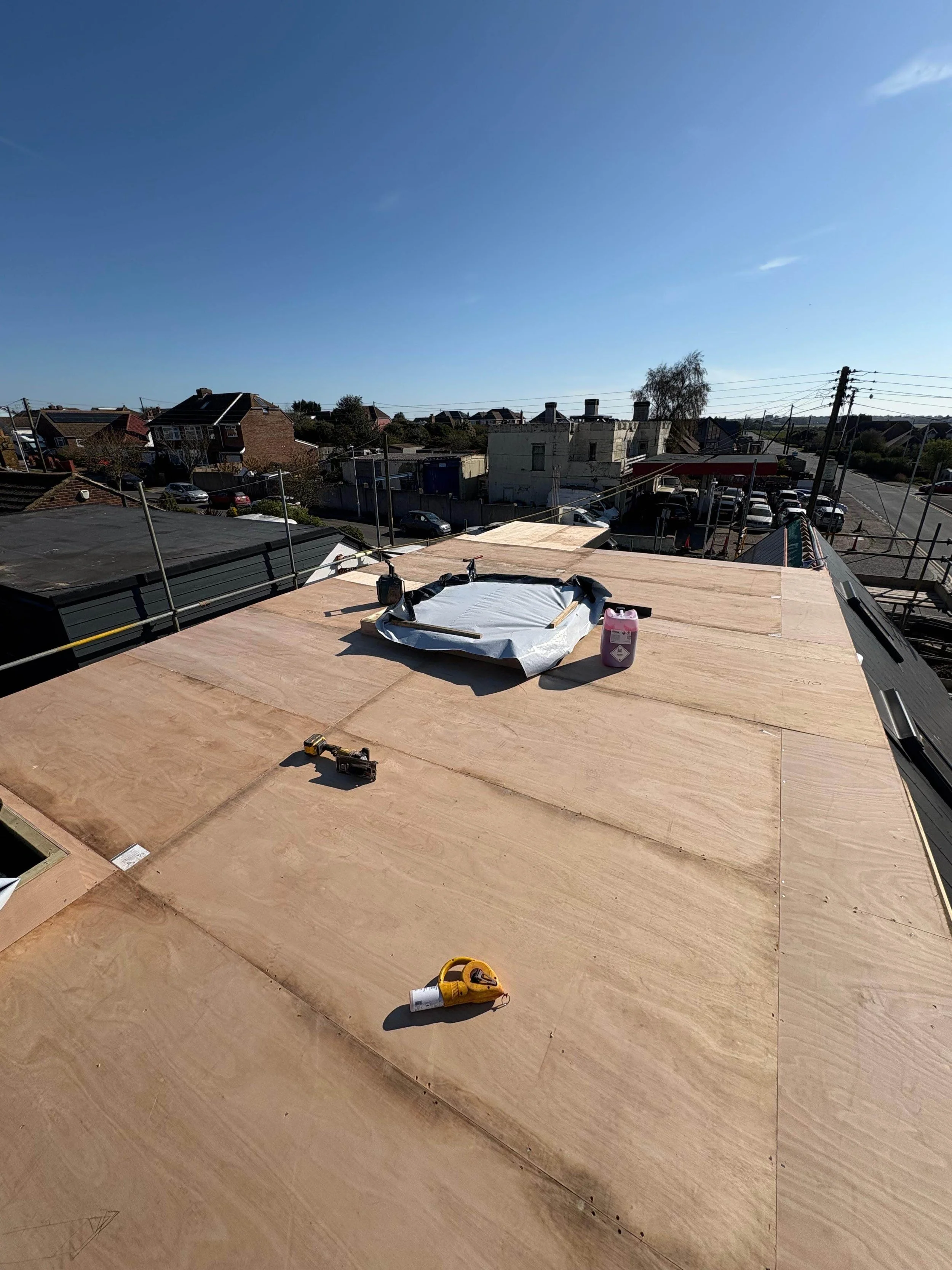 Roof under construction with plywood sheets, construction tools, and materials on top, with a clear blue sky and neighboring buildings in the background.