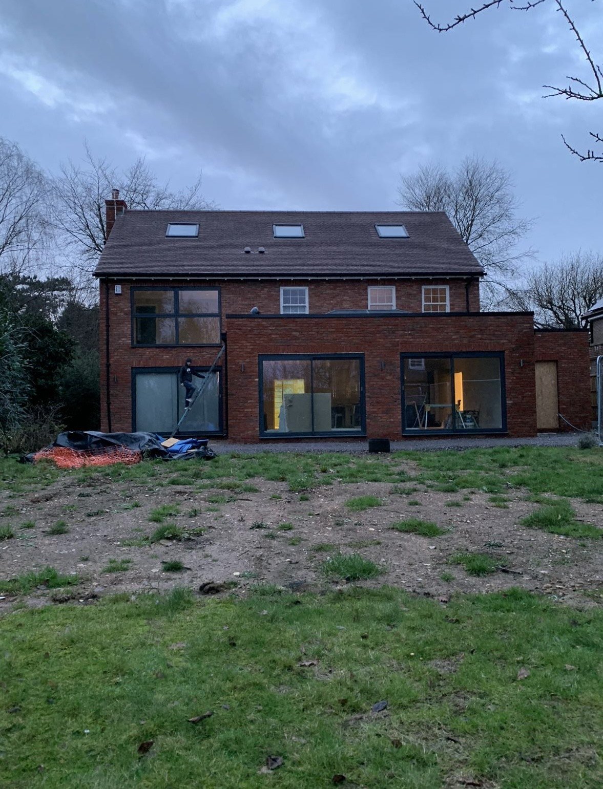 Back of a brick house under construction, with large glass sliding doors, a person working on the side, and unfinished yard with patches of dirt and grass.