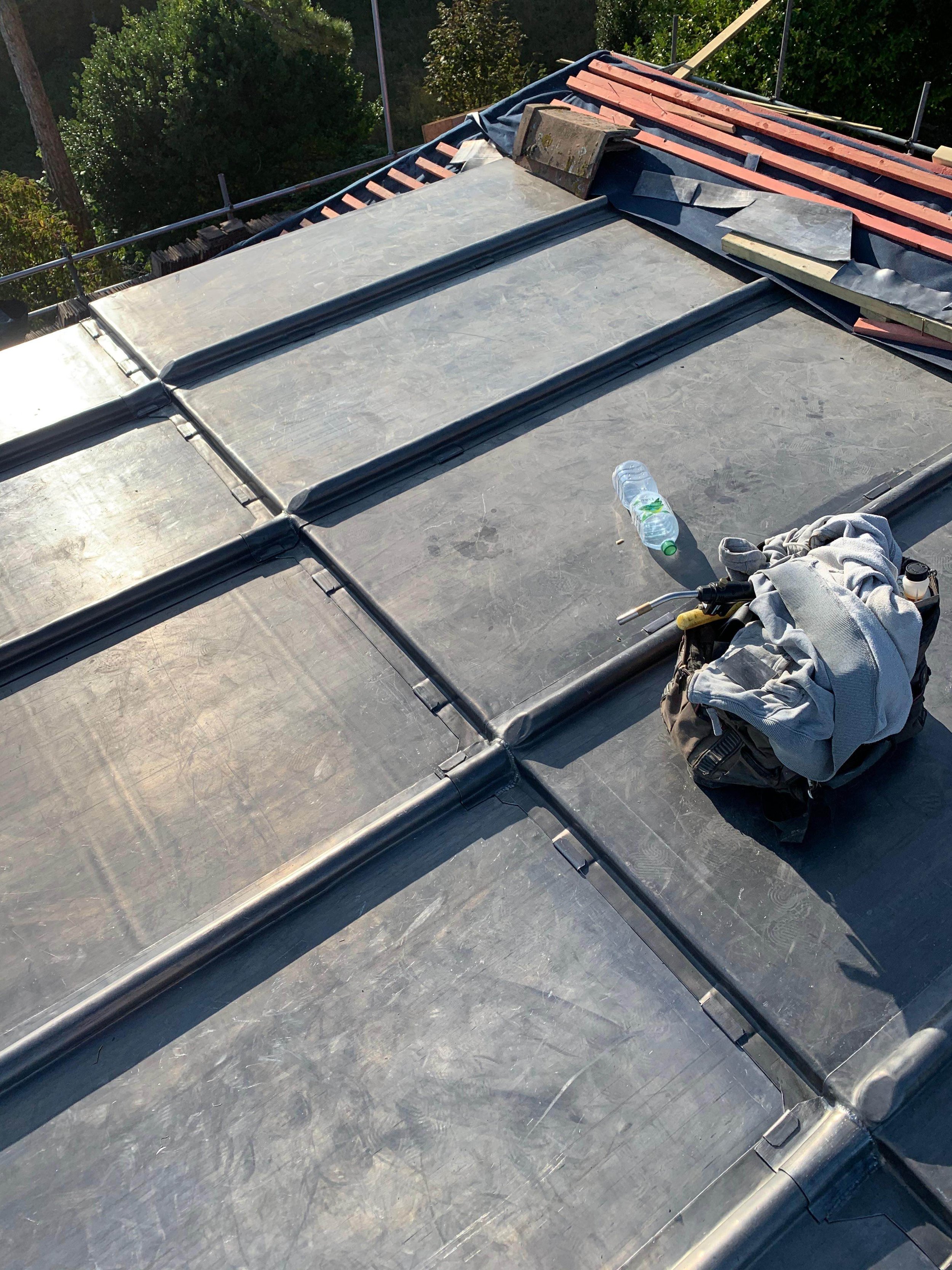 A rooftop under construction with black roofing material, a water bottle, a work bag with tools, and insulation piles, surrounded by green trees and scaffolding.