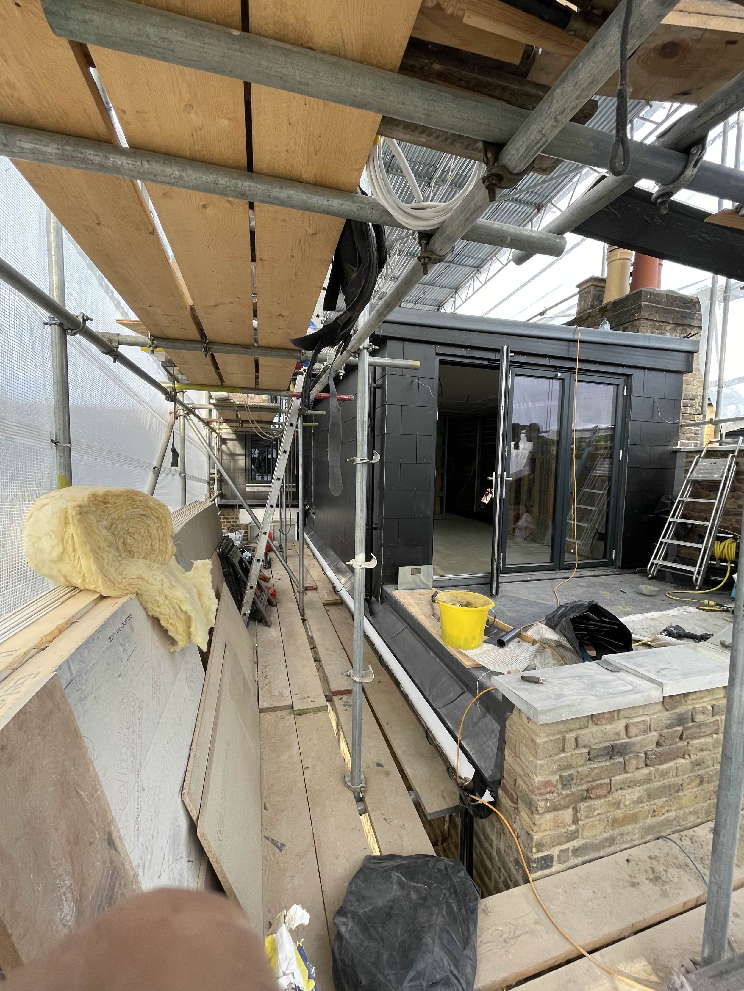 Construction site with scaffolding, partially built brick and black siding exterior wall, glass door, construction tools, and materials.