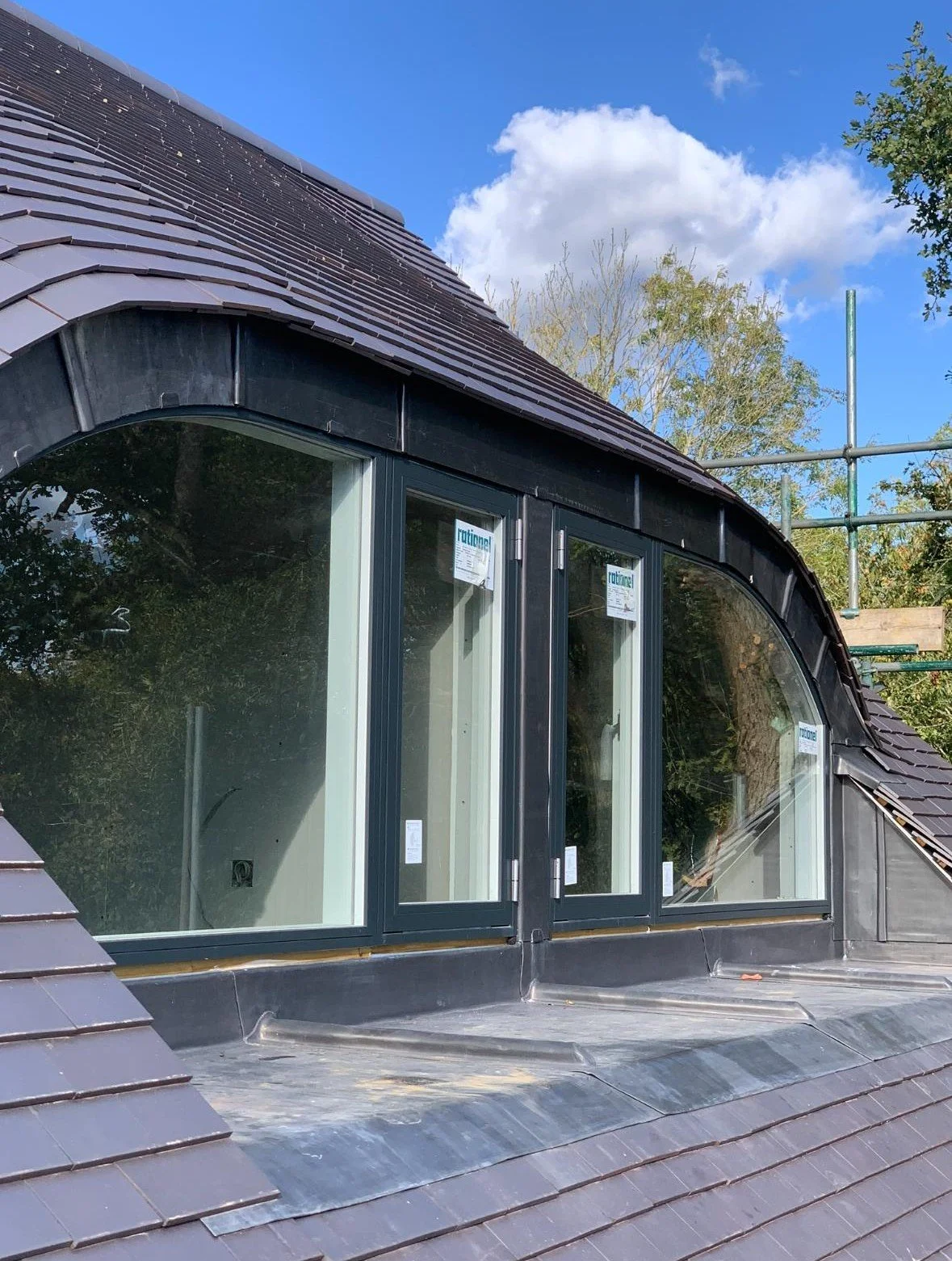 View of a roof with new climate windows installed in a building under construction, with scaffolding visible and trees in the background.