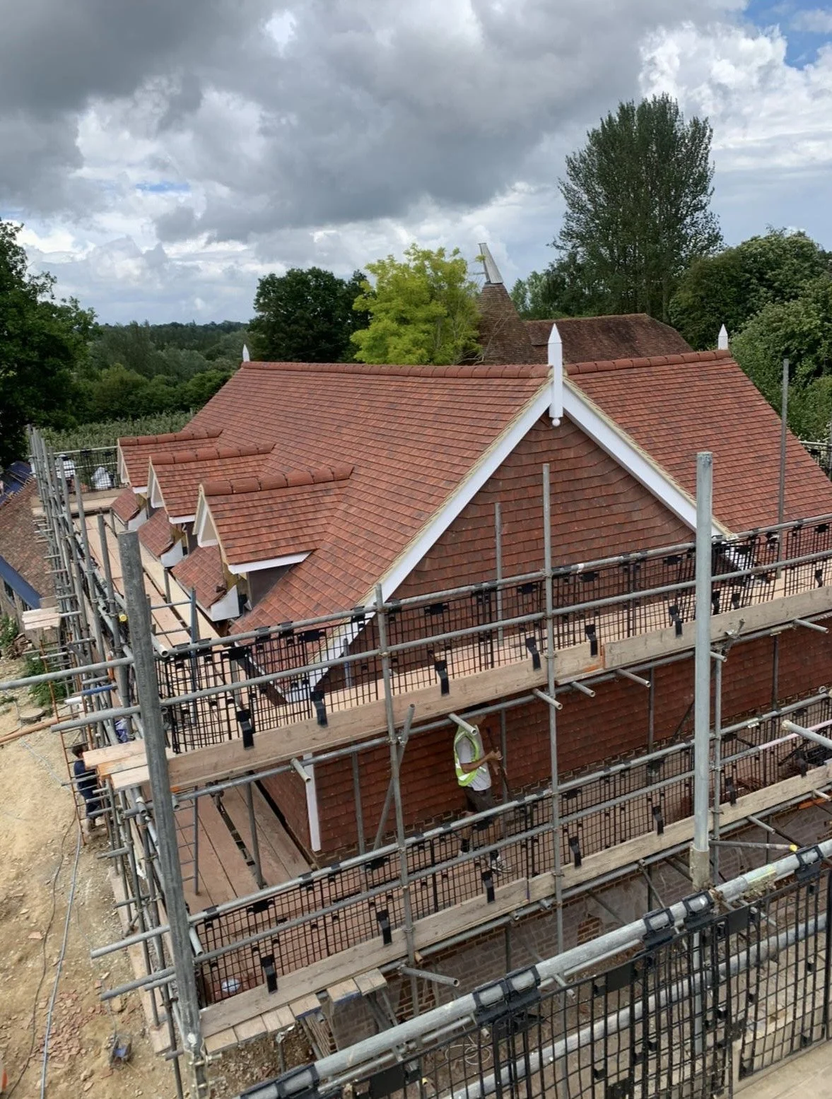Construction workers on scaffolding working on the roof of a building with red tiles, surrounded by trees and cloudy sky