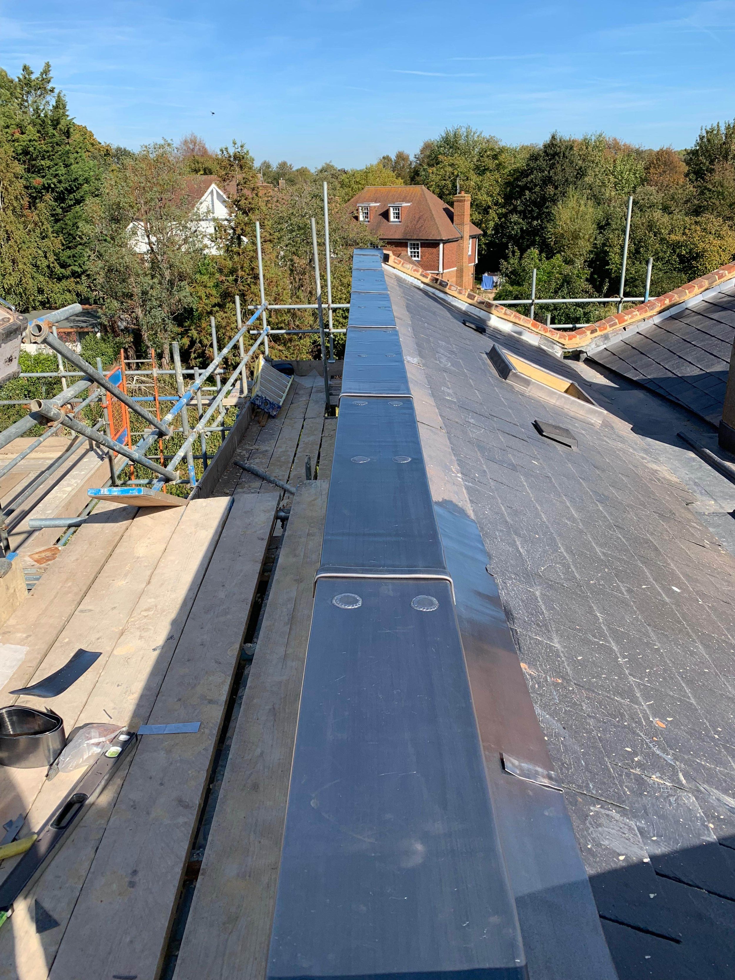 View of a house roof under construction with scaffolding around it, showing roofing materials and tools, and a background of trees and neighboring houses under a clear blue sky.