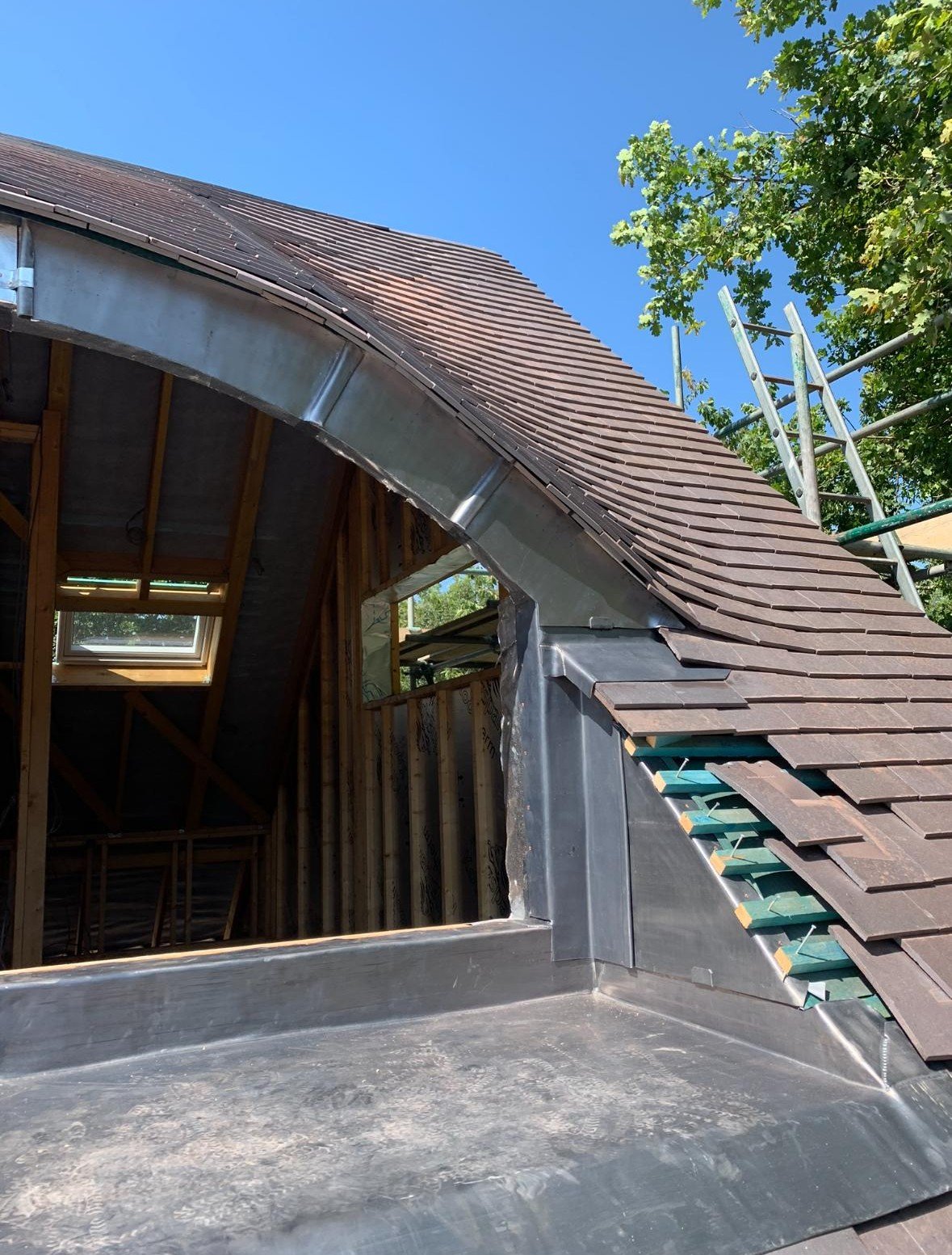Construction site showing a partially installed roof with brown shingles, exposed wooden framing inside, and a metal flashing around the edge. A ladder is visible on the right, and a tree with green leaves is in the background under a clear blue sky.
