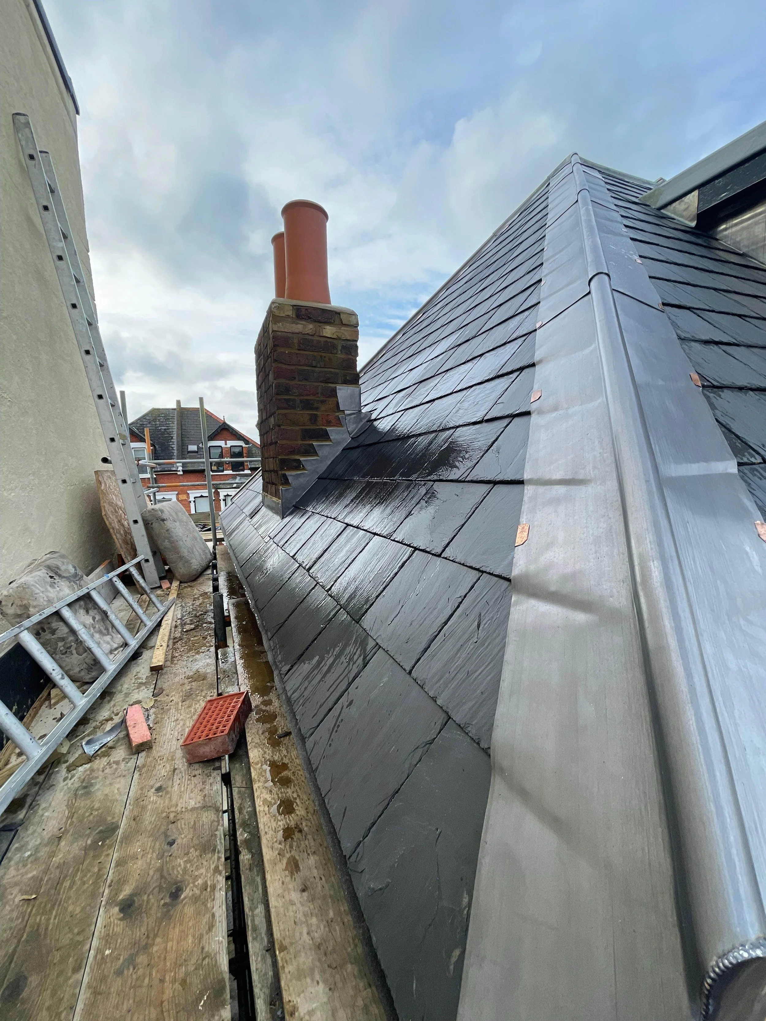 Close-up of a newly installed black slate roof with a metal edge, a brick chimney, and three red clay chimney pipes against a partly cloudy sky.