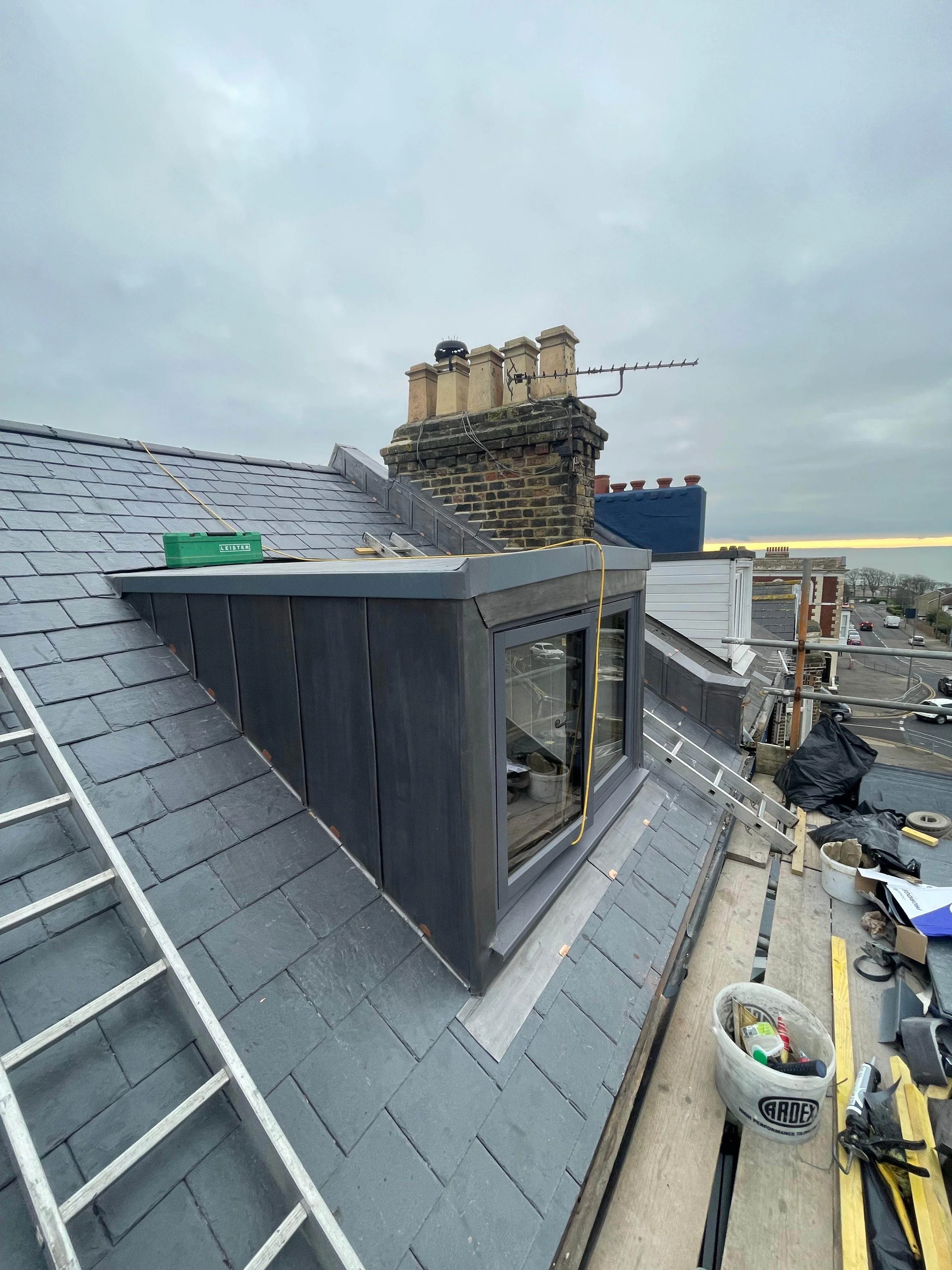 Construction site on a sloped roof with tools and materials, including a ladder, black plastic covering, and a new window installation.