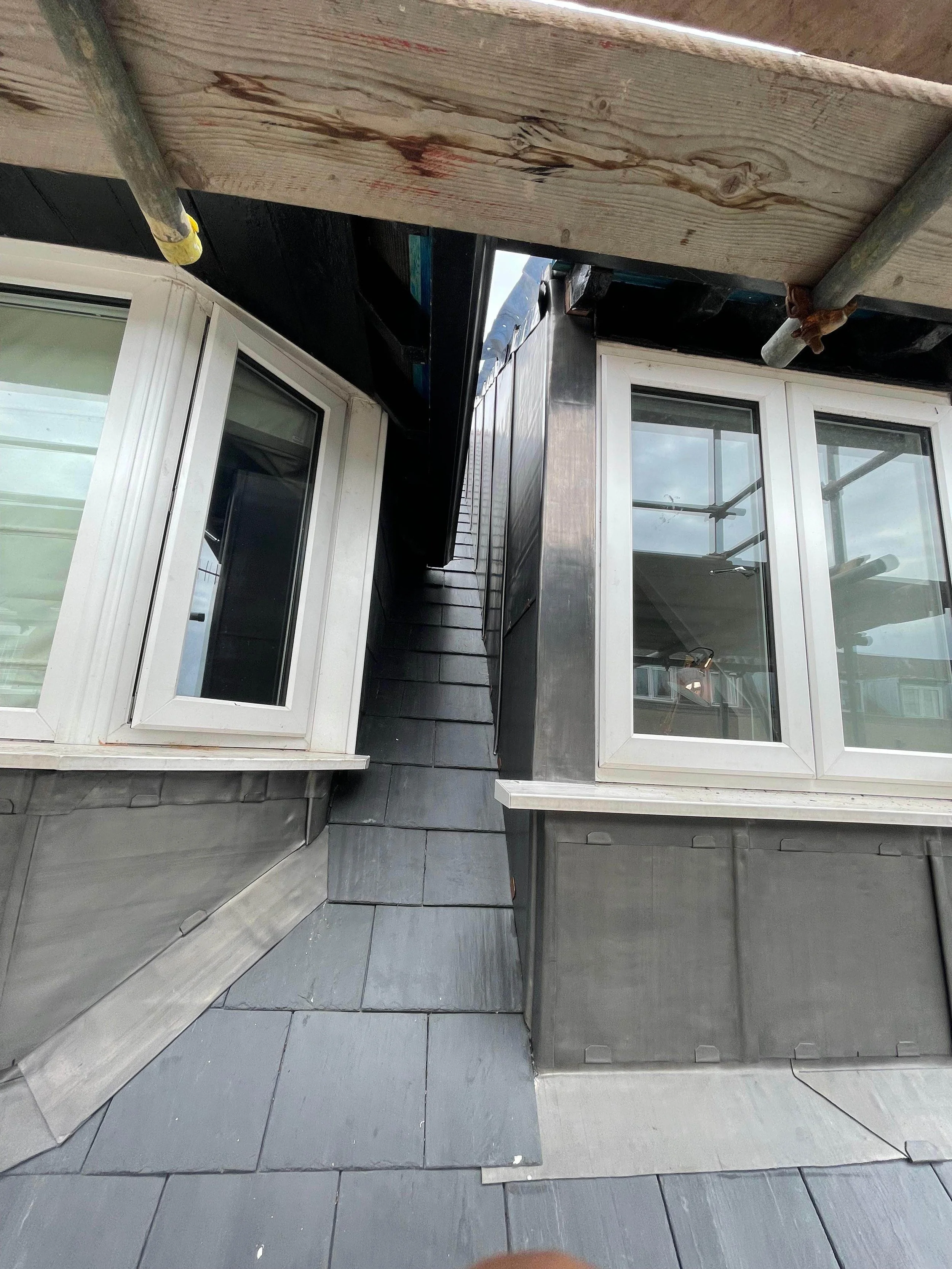 View looking up at the roof of a building from a narrow ledge with windows on both sides and scaffolding above.