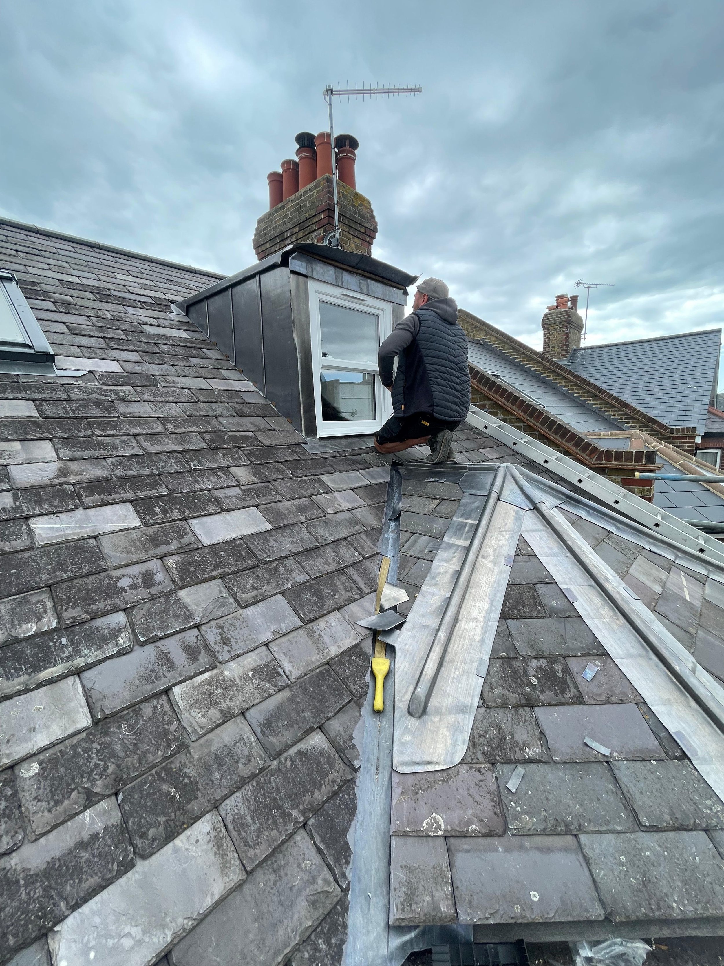 A man working on a sloped rooftop with curved tiles, using tools, with a chimney and a small dormer window nearby, set against a cloudy sky.
