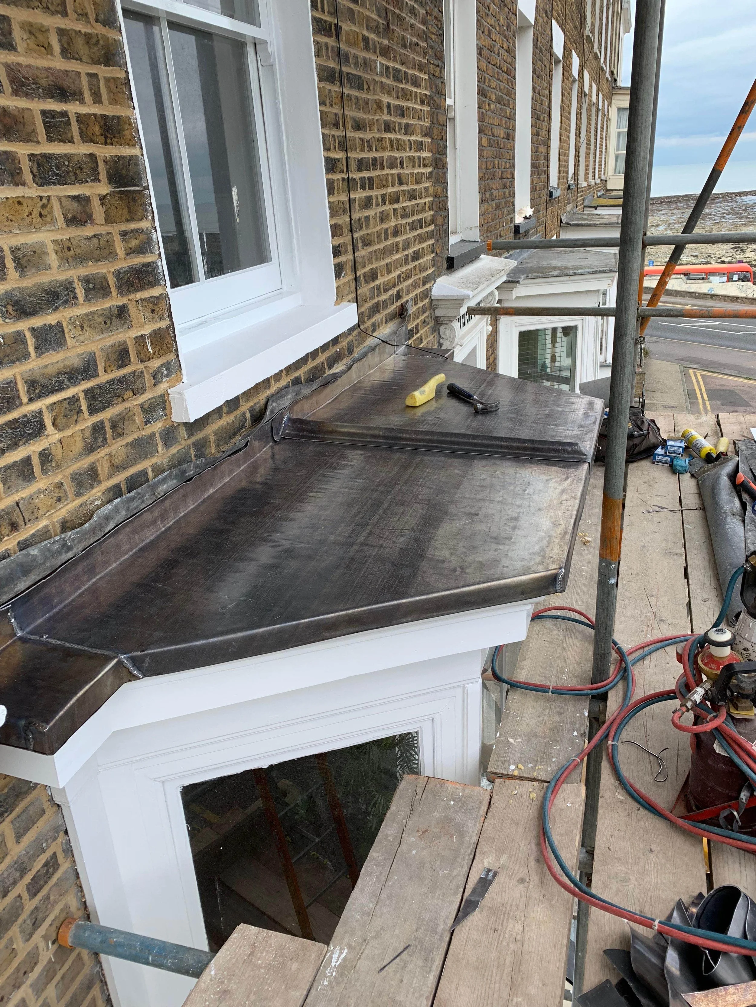 Construction workers installing a metal roof for a window on a brick building, with construction tools and equipment on scaffolding.